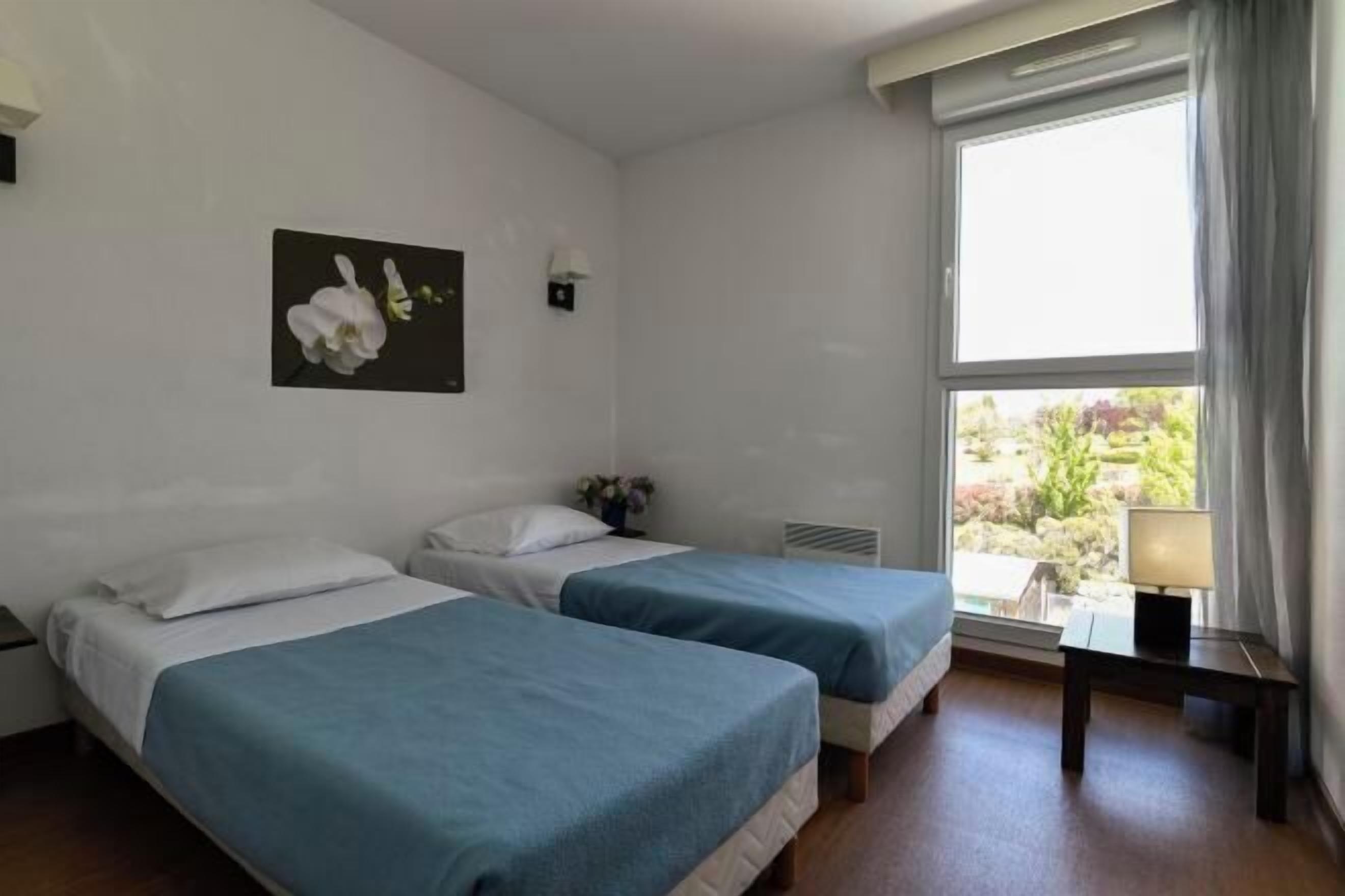 Habitación de hotel con dos camas, una ventana con vista a follaje, una mesita con lámpara y un cuadro colgado en la pared de una orquídea.