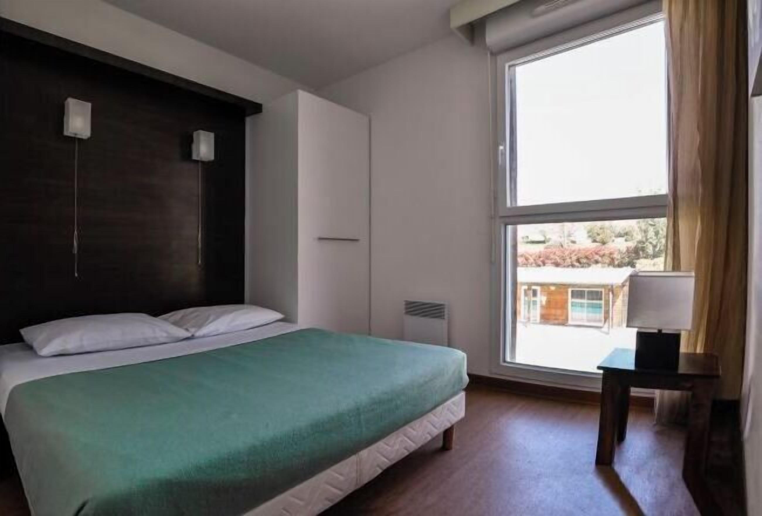 Un dormitorio con un ventanal grande, una cama con colcha verde, una mesita de noche con lámpara y un cabecero de madera oscura.