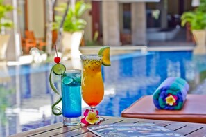 Two cocktails on a table by a poolside with a towel and a flower.