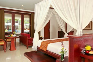 A hotel room with a bed, a sitting area with a table and chairs, a small table with a vase of flowers, and a view of the outside. - The Rani Hotel & Spa (Kuta)