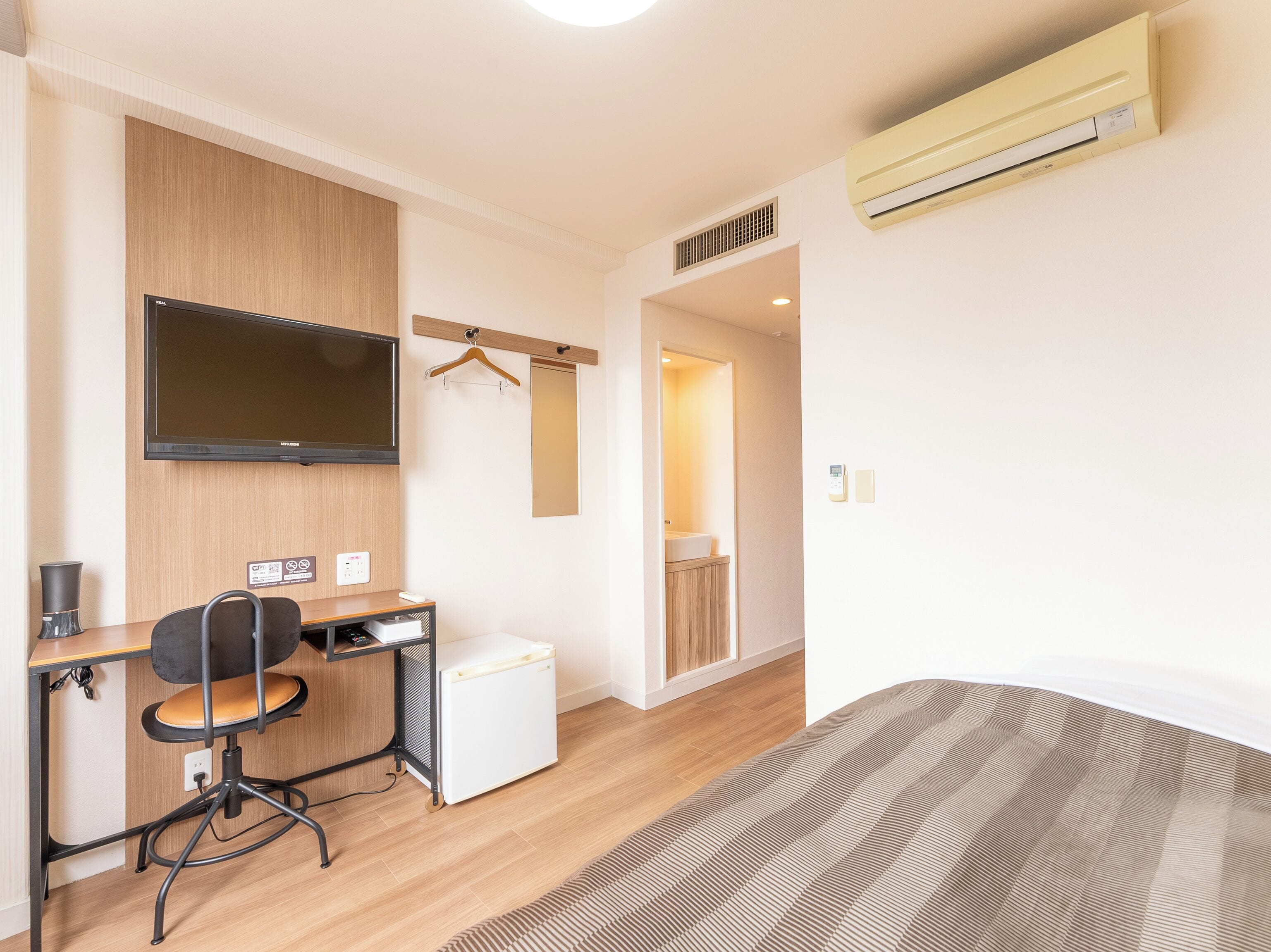 Quarto de hotel com escrivaninha de madeira, cadeira, televisão, mini-geladeira, cama e banheiro visível através de uma porta aberta.