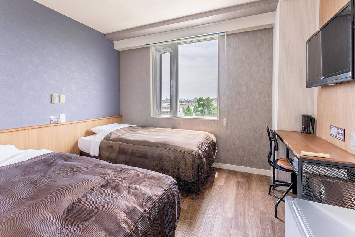 Habitación de hotel con dos camas, un escritorio, una silla, un televisor y una ventana con vista a la ciudad.