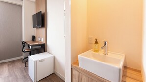 Une pièce de taille modeste, dotée d’un bureau en bois, d’un petit réfrigérateur, d’un téléviseur fixé au mur et d’un lavabo blanc avec un distributeur de savon.