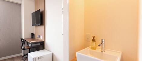 A compact room with a wooden desk, a small refrigerator, a television mounted on the wall, and a white sink with a soap dispenser.