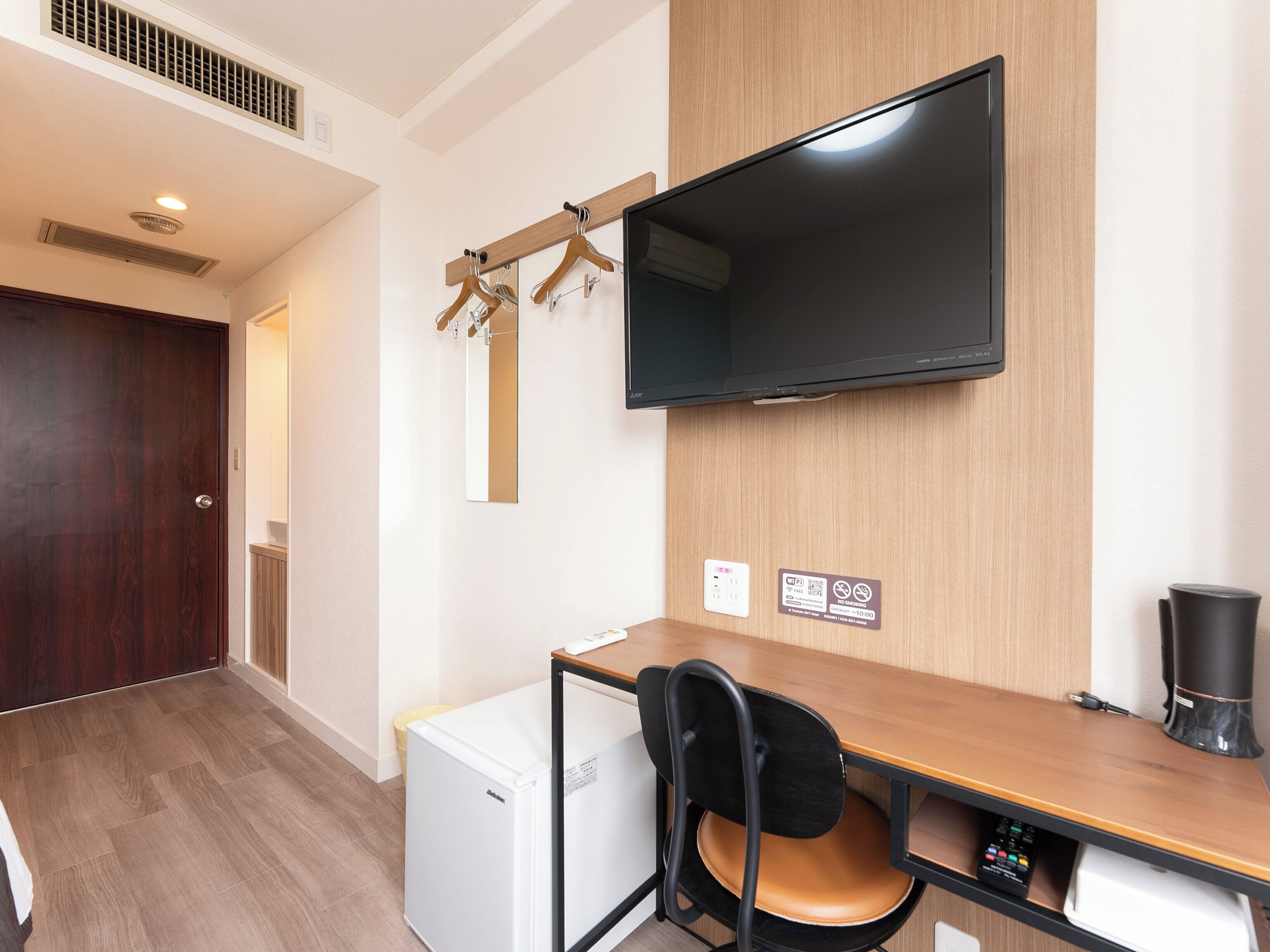 A compact hotel room with a wooden desk, a white mini-fridge, a black chair, a mounted TV, and a wooden door.