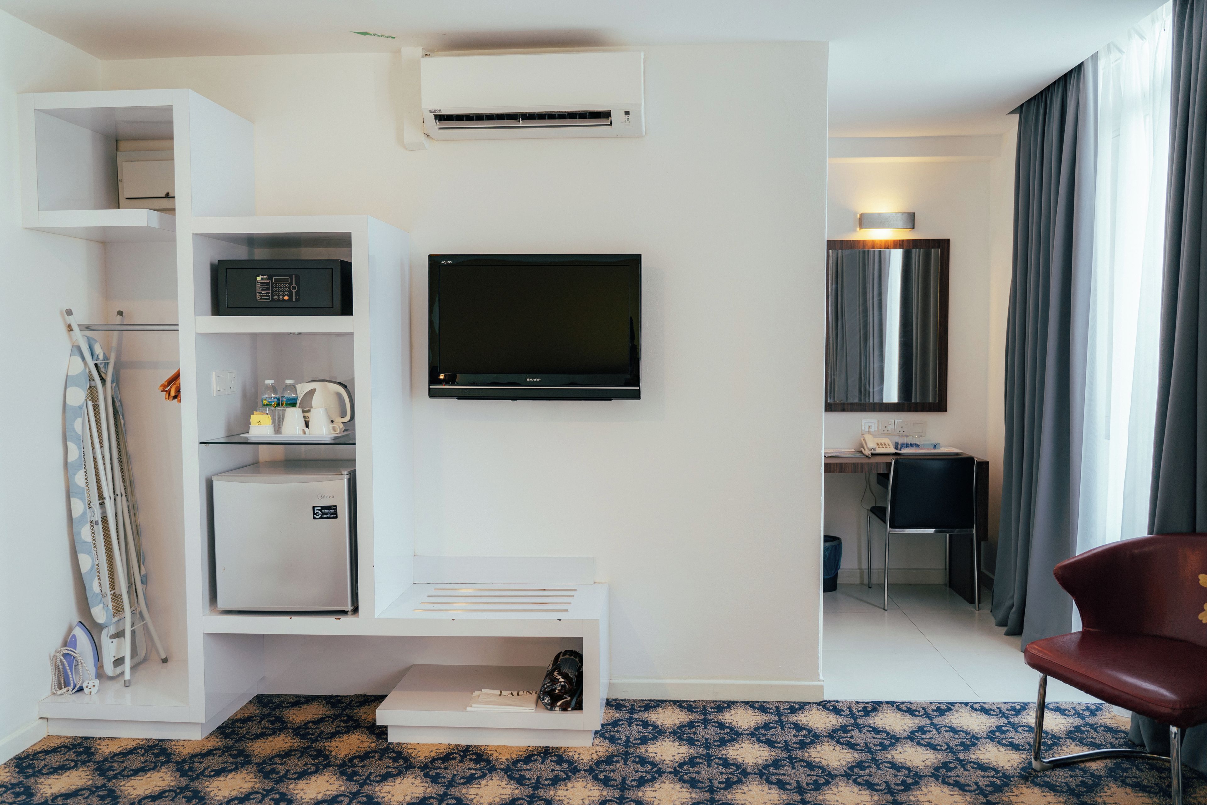 A hotel room with a flat-screen TV mounted on the wall, a mini-fridge, a microwave, a small seating area with a chair, and a desk with a lamp.