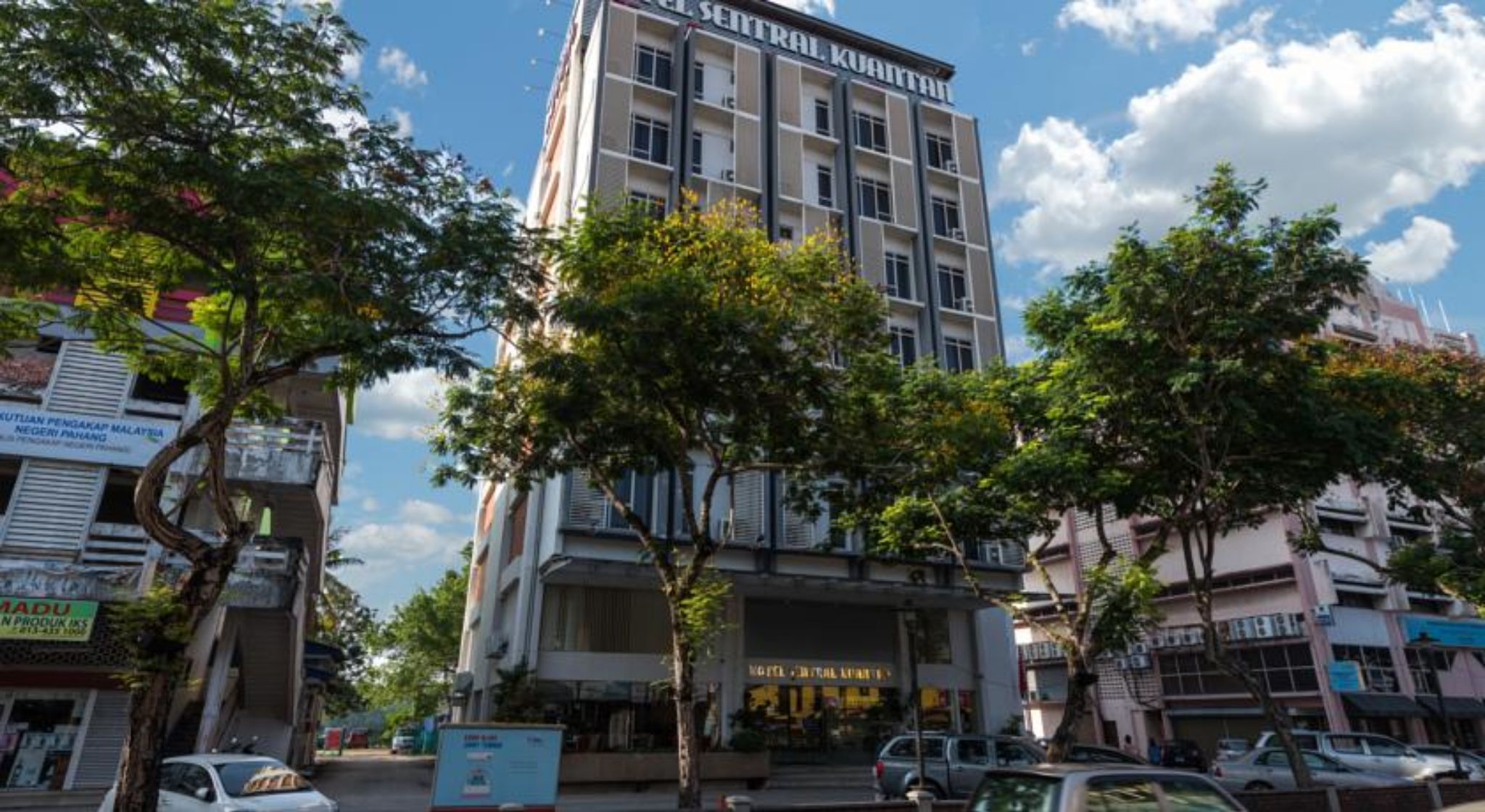 Un hotel a più piani con un ingresso centrale, circondato da alberi e altri edifici.