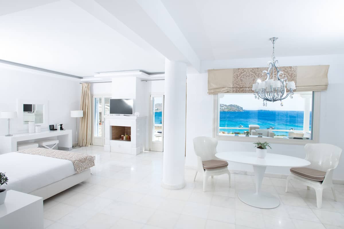 A modern hotel room with a large bed, a dining area, a TV, and a view of the sea.