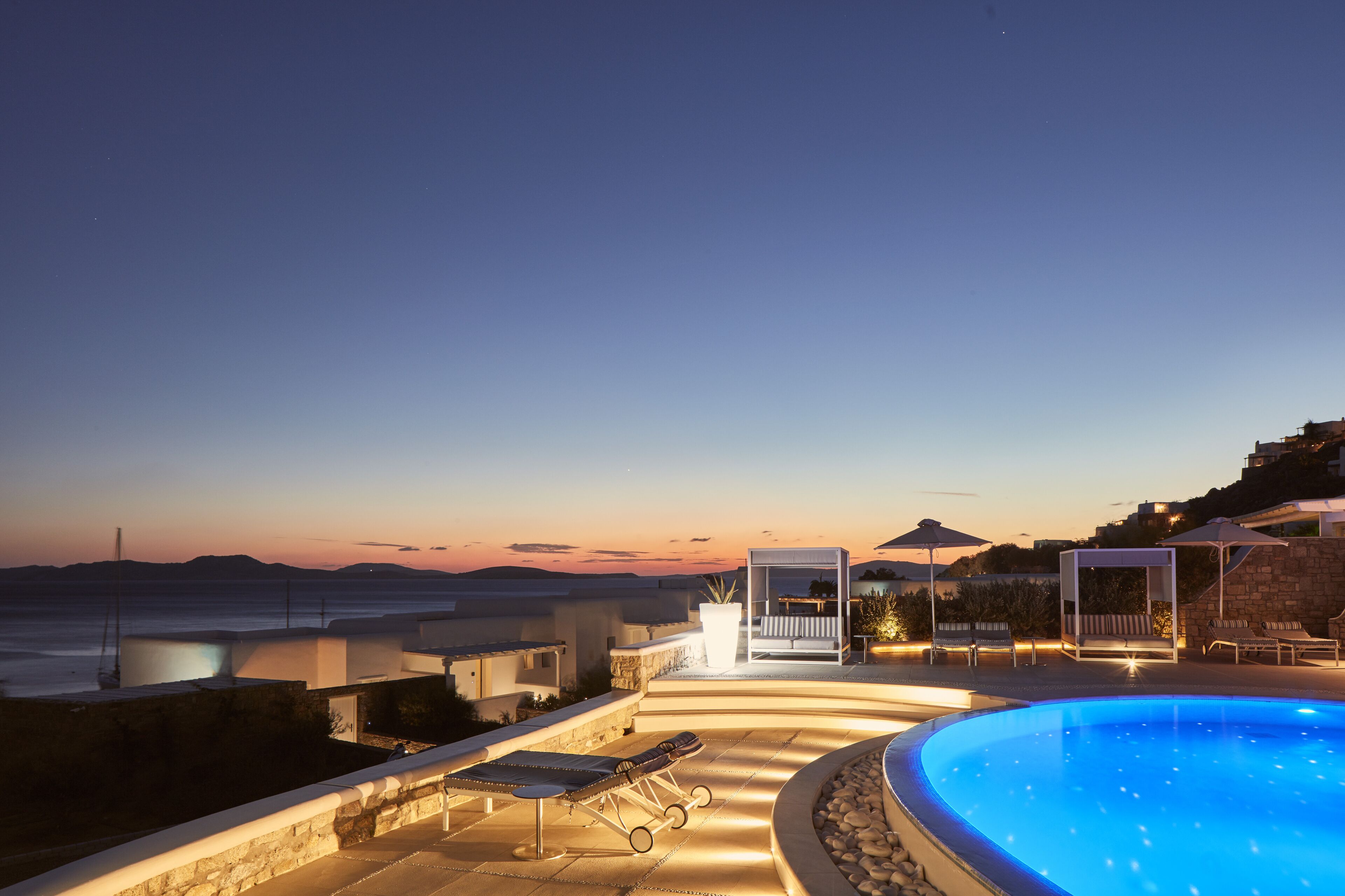 A pool area with lounge chairs and umbrellas, overlooking a scenic ocean view at dusk.