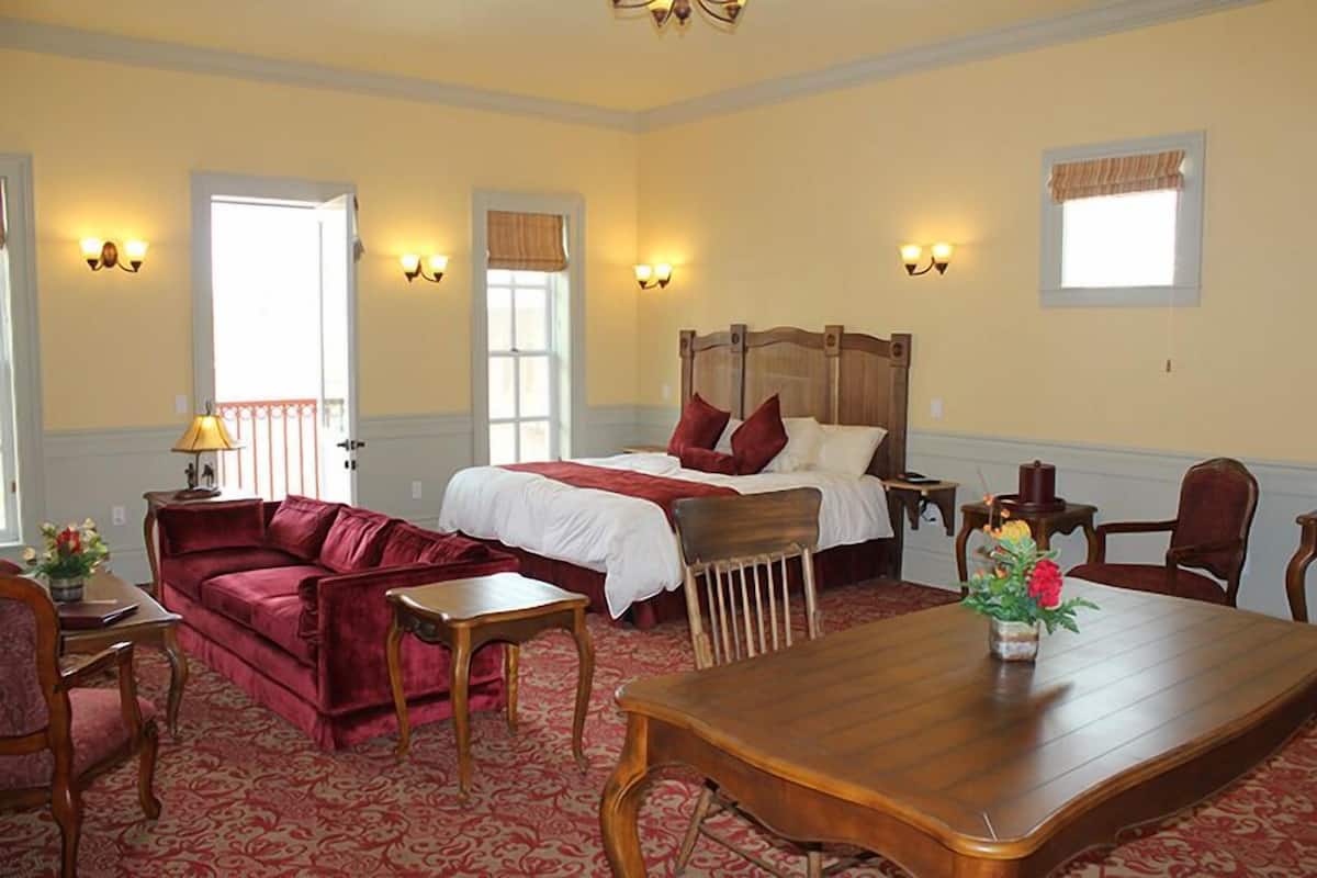 A hotel room with a bed, a red velvet sofa, a wooden dining table, and a small table with a lamp.