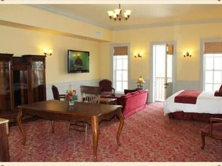 A hotel room with a wooden dining table, a sofa, a bed with a red blanket, and a chandelier.