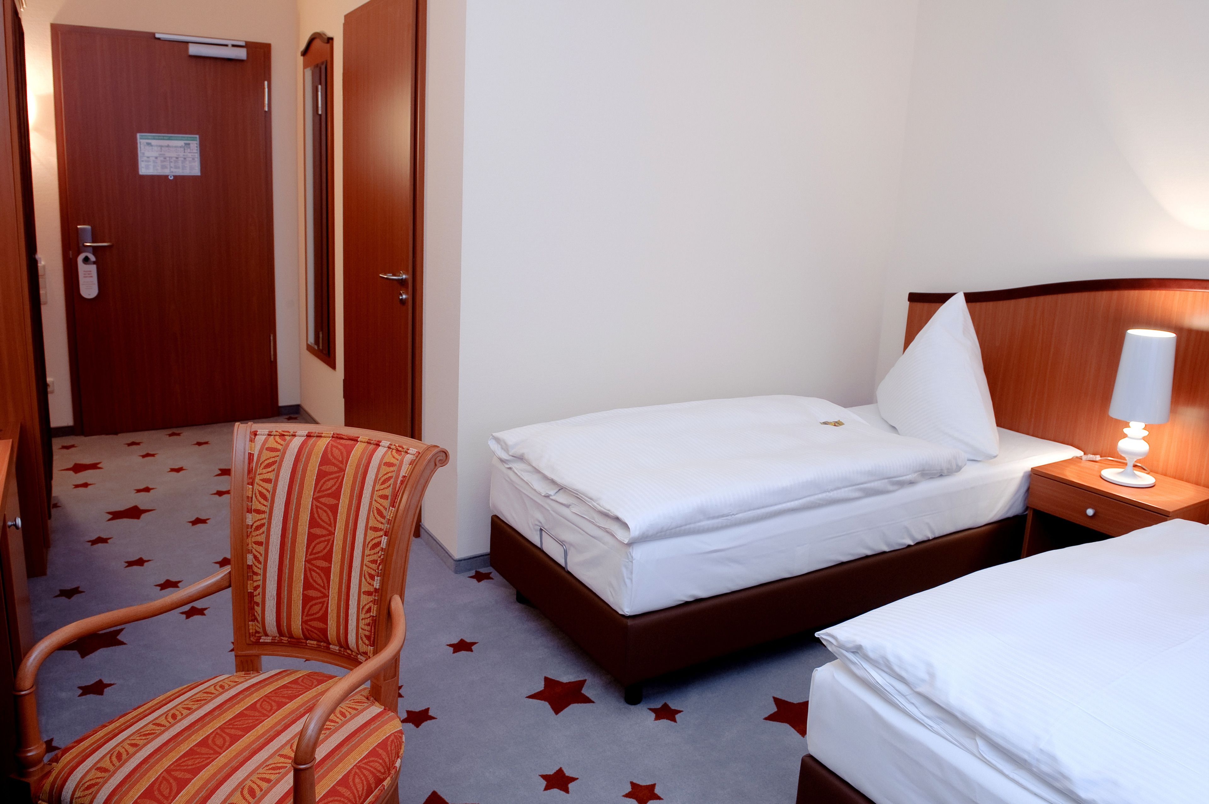 Habitación de hotel con cama, una silla, una mesita de noche con lámpara y una puerta con un cartel.