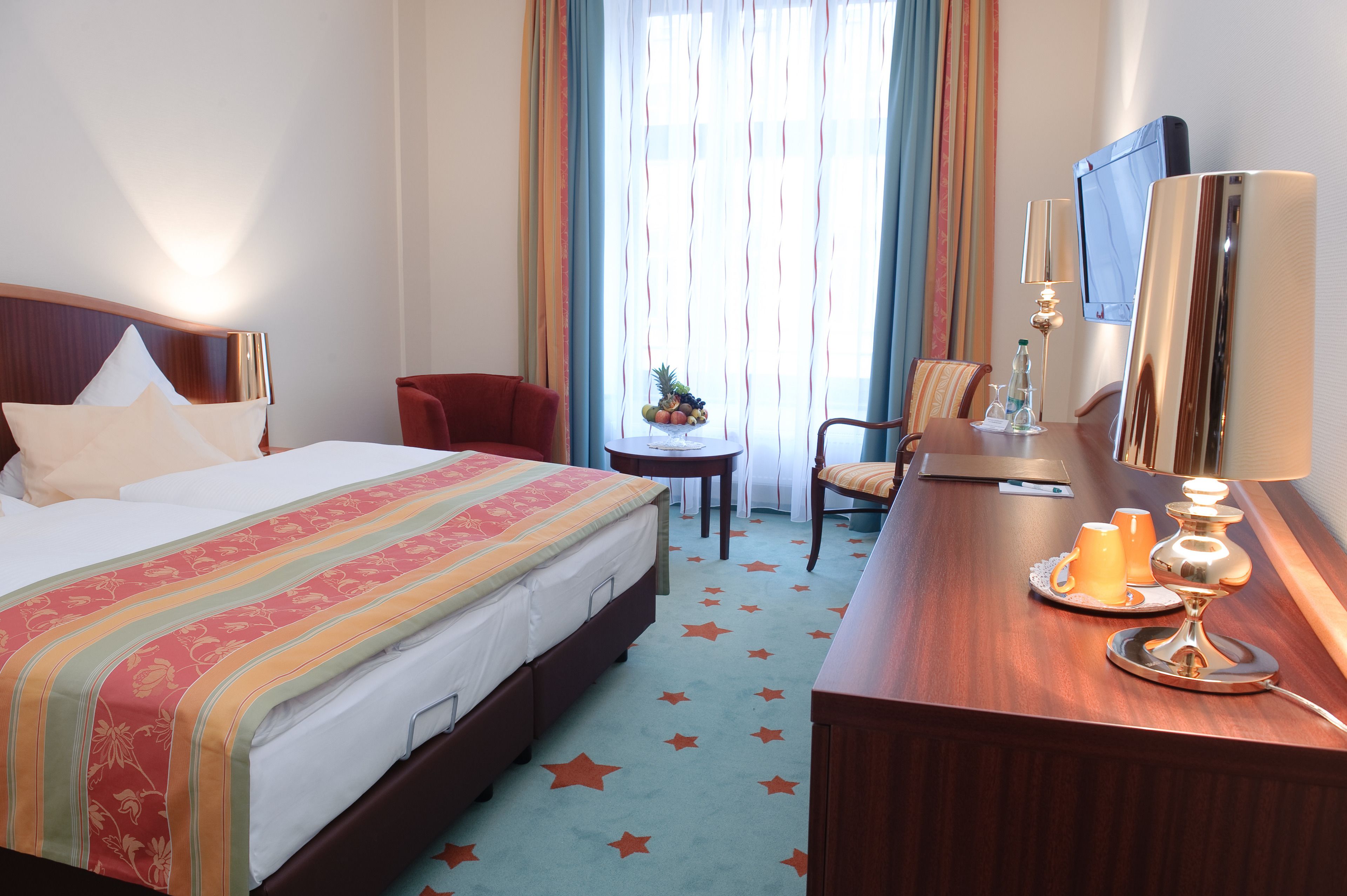 A hotel room with a bed, a desk with a lamp, a chair, a small table with fruit, and a TV.