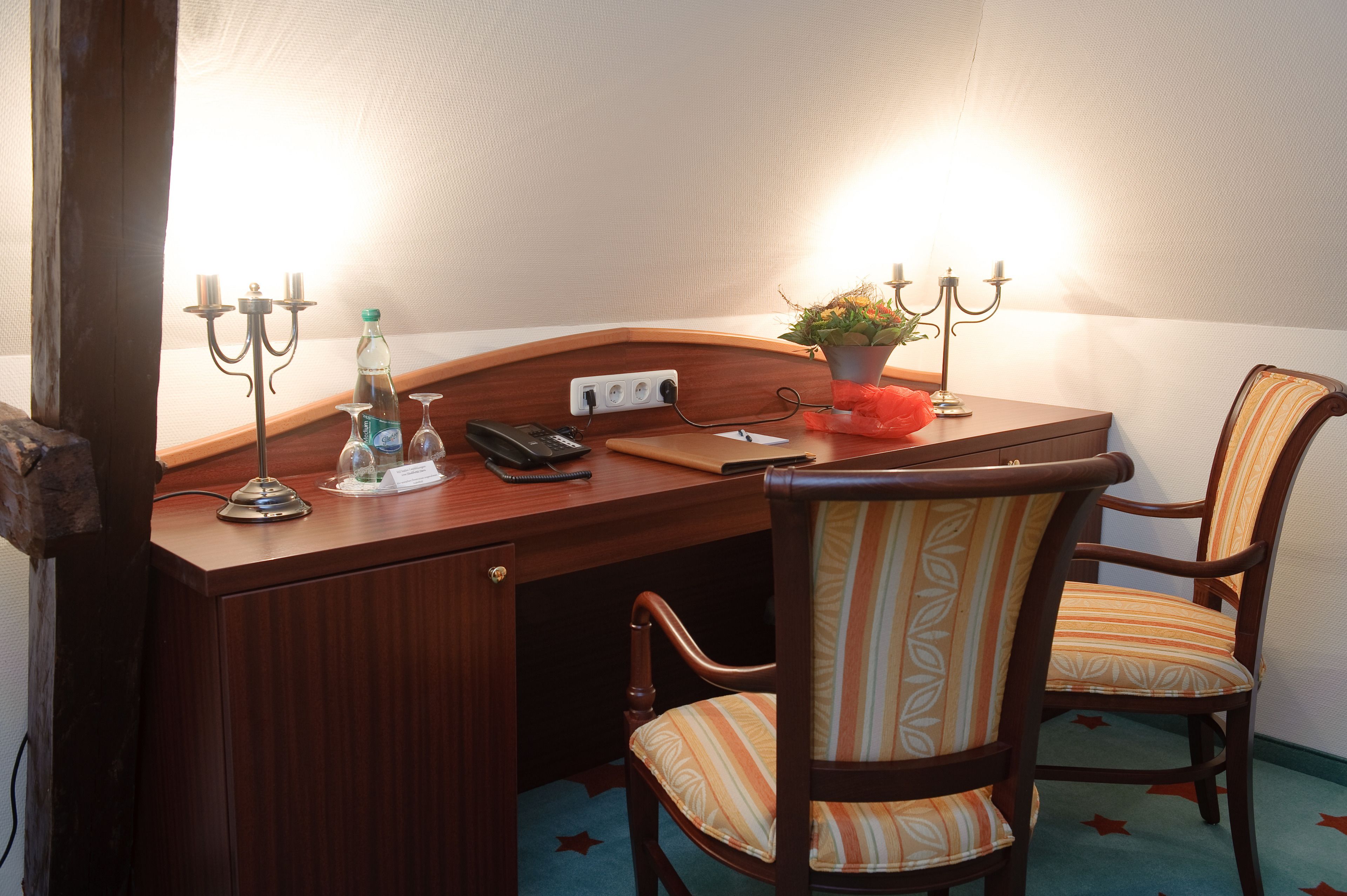 Un bureau en bois avec un téléphone, une lampe, une plante et un chandelier. On y trouve deux chaises à rayures et une petite table avec une bouteille et des verres.