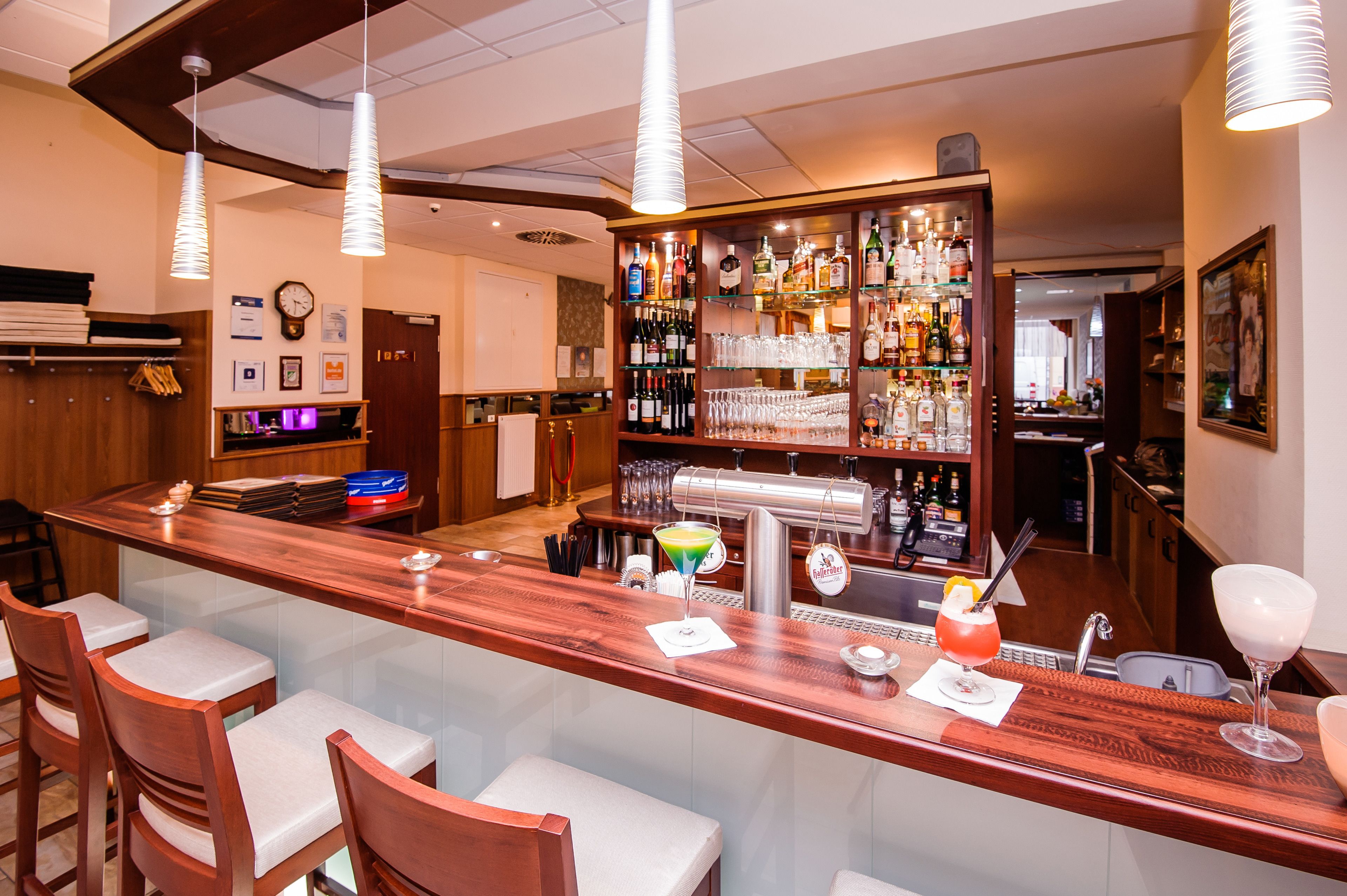 Uma área de bar com balcão de madeira, vários bancos e uma variedade de garrafas e copos.