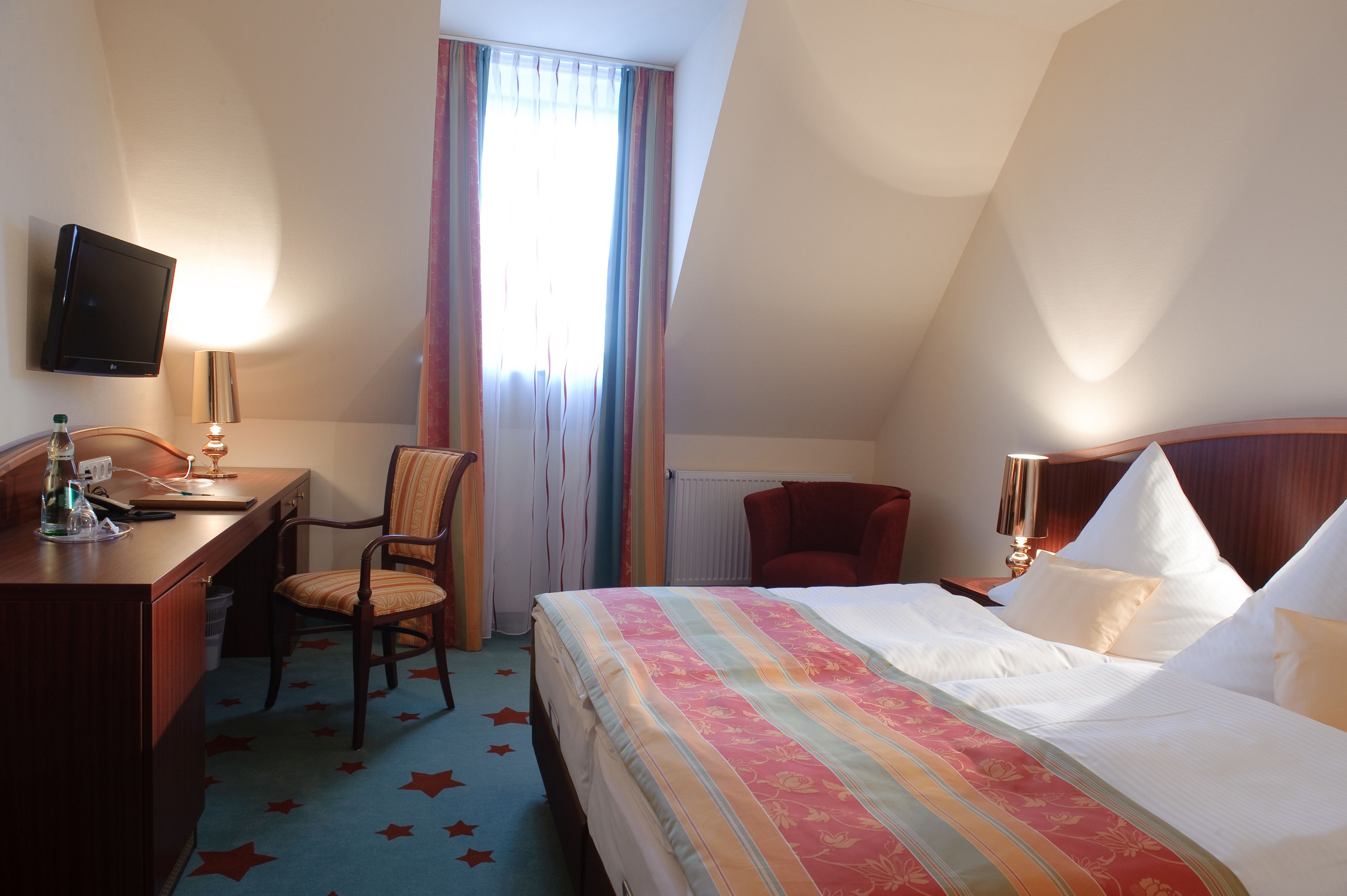 A hotel room with a bed, desk, chair, TV, and curtains.