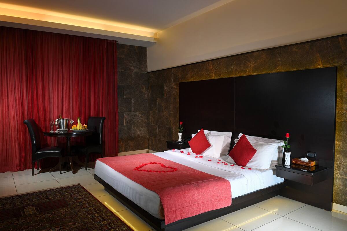 Une chambre d’hôtel avec un grand lit, des rideaux rouges, une petite table avec un vase et des fleurs, et une tête de lit sombre.