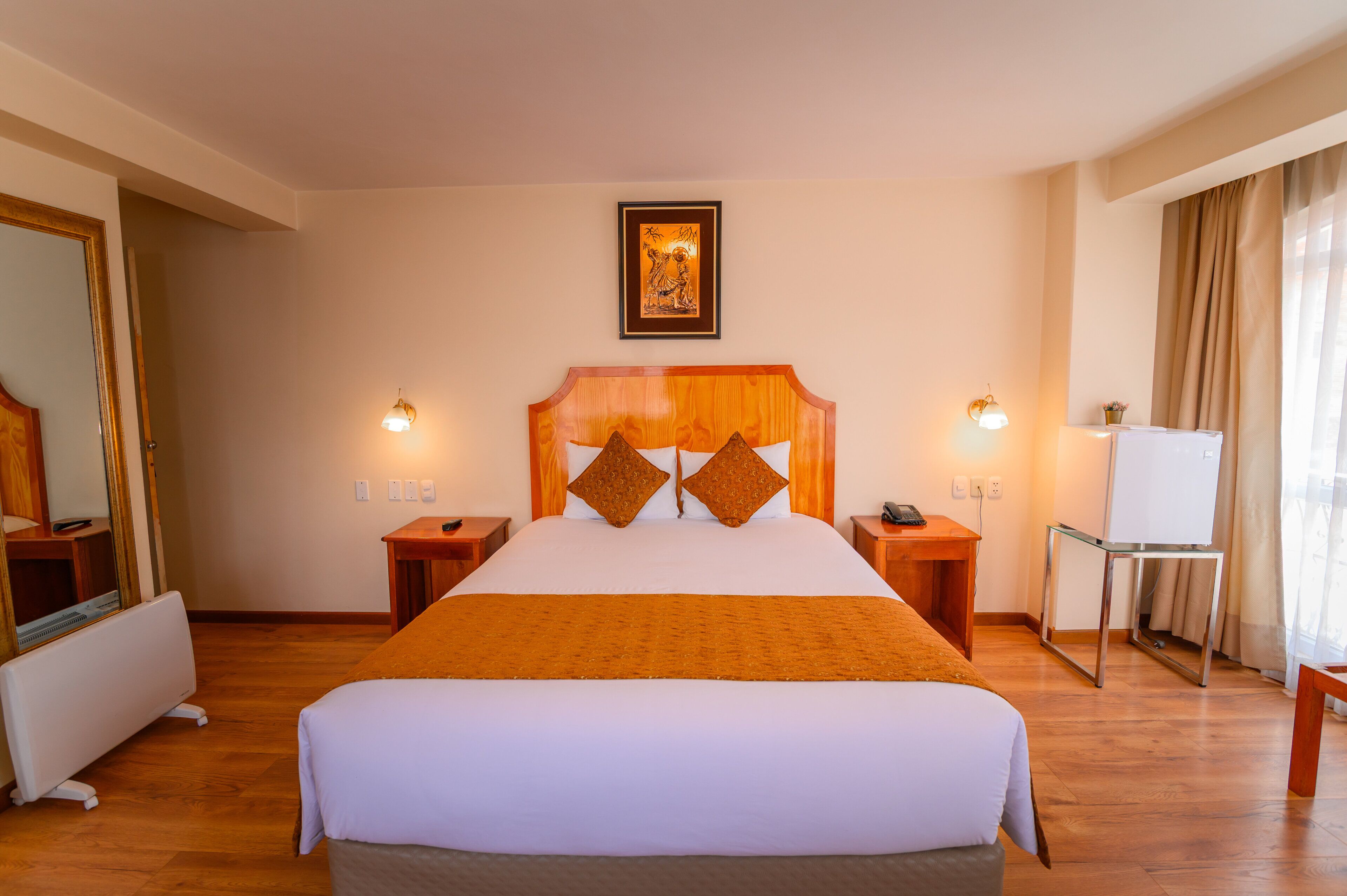 A hotel room with a large bed, wooden headboard, bedside tables, a desk, a chair, a mirror, and a framed picture on the wall.