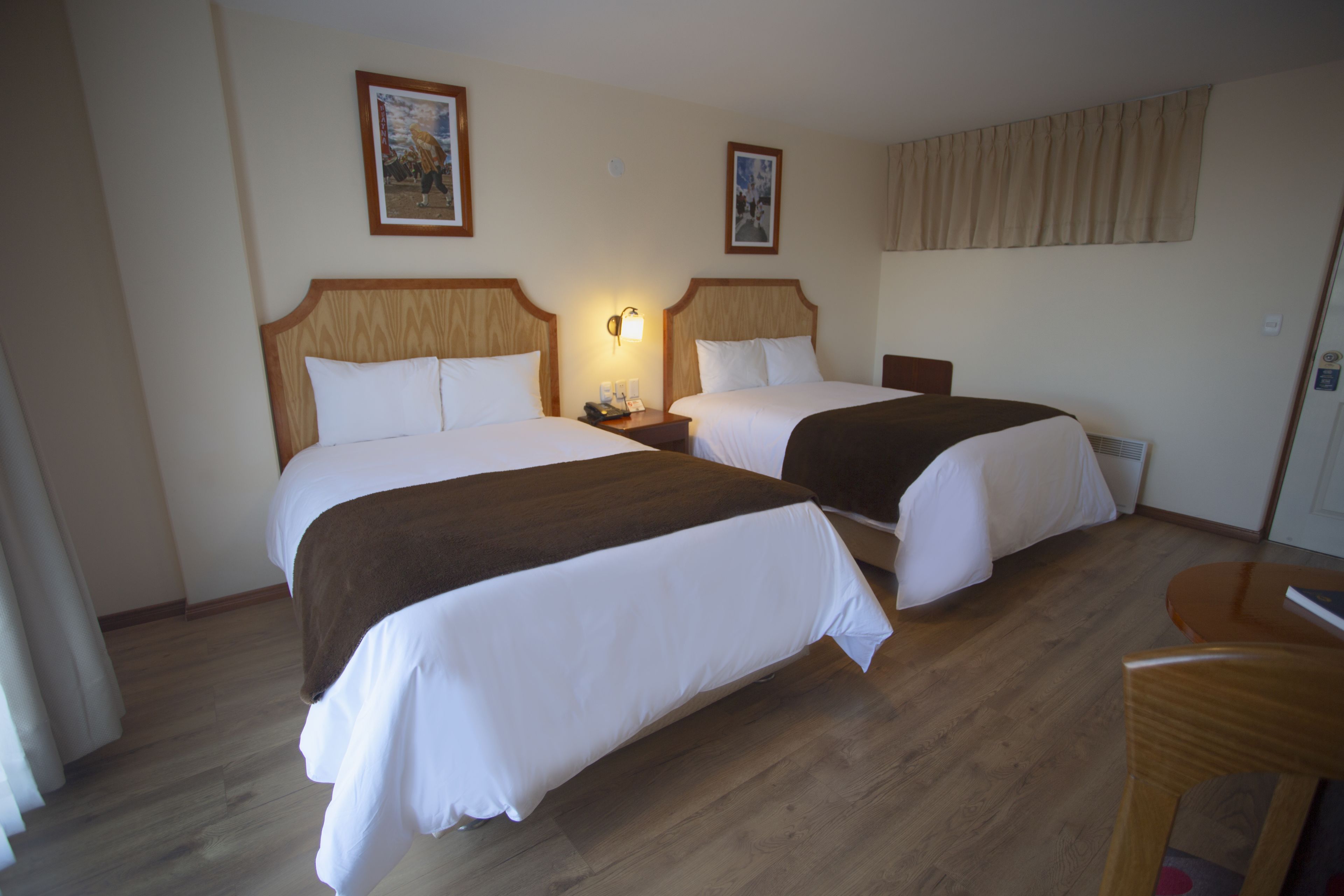 A hotel room with two beds, wooden headboards, white bedding, a nightstand with a lamp, and a wooden desk.