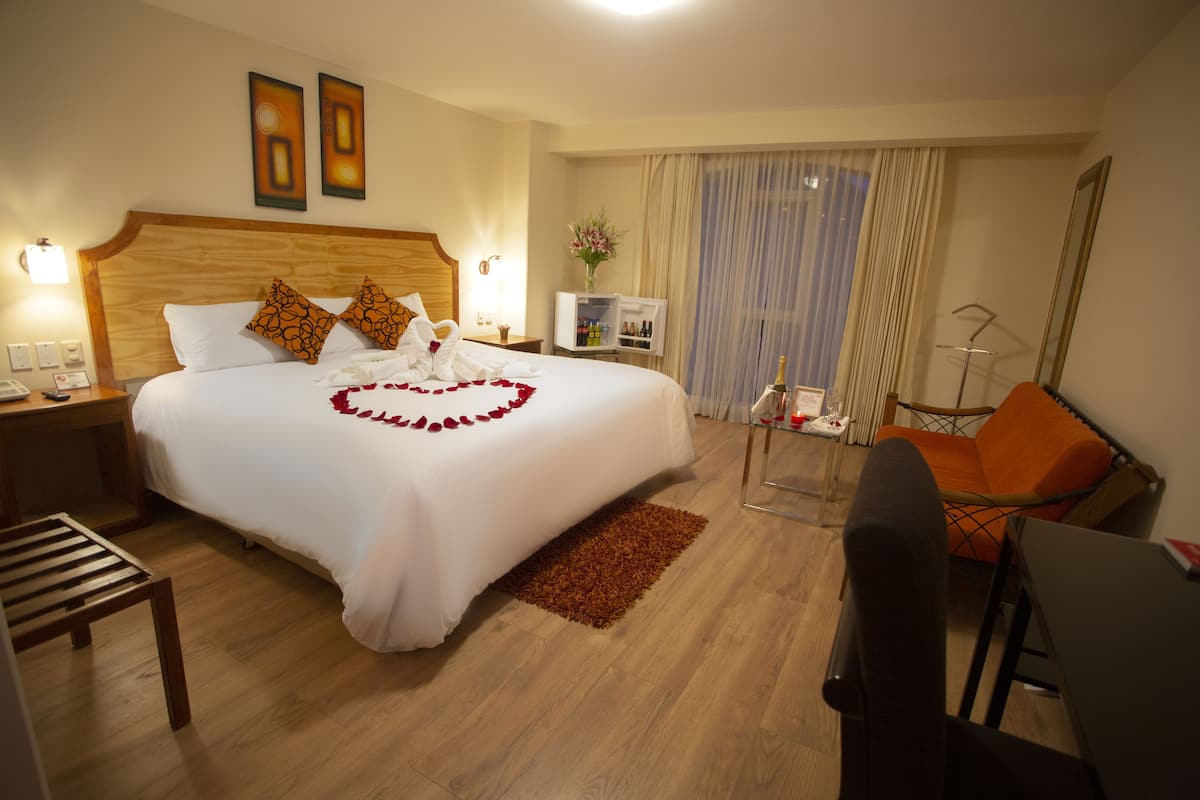 Habitación de hotel con una cama grande, un escritorio, una silla naranja, una mesita pequeña, una mesita de noche, una lámpara, un espejo, una ventana con cortinas y un televisor instalado en la pared.