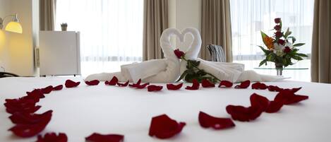 A hotel room with a bed adorned with rose petals and a heart-shaped towel arrangement.