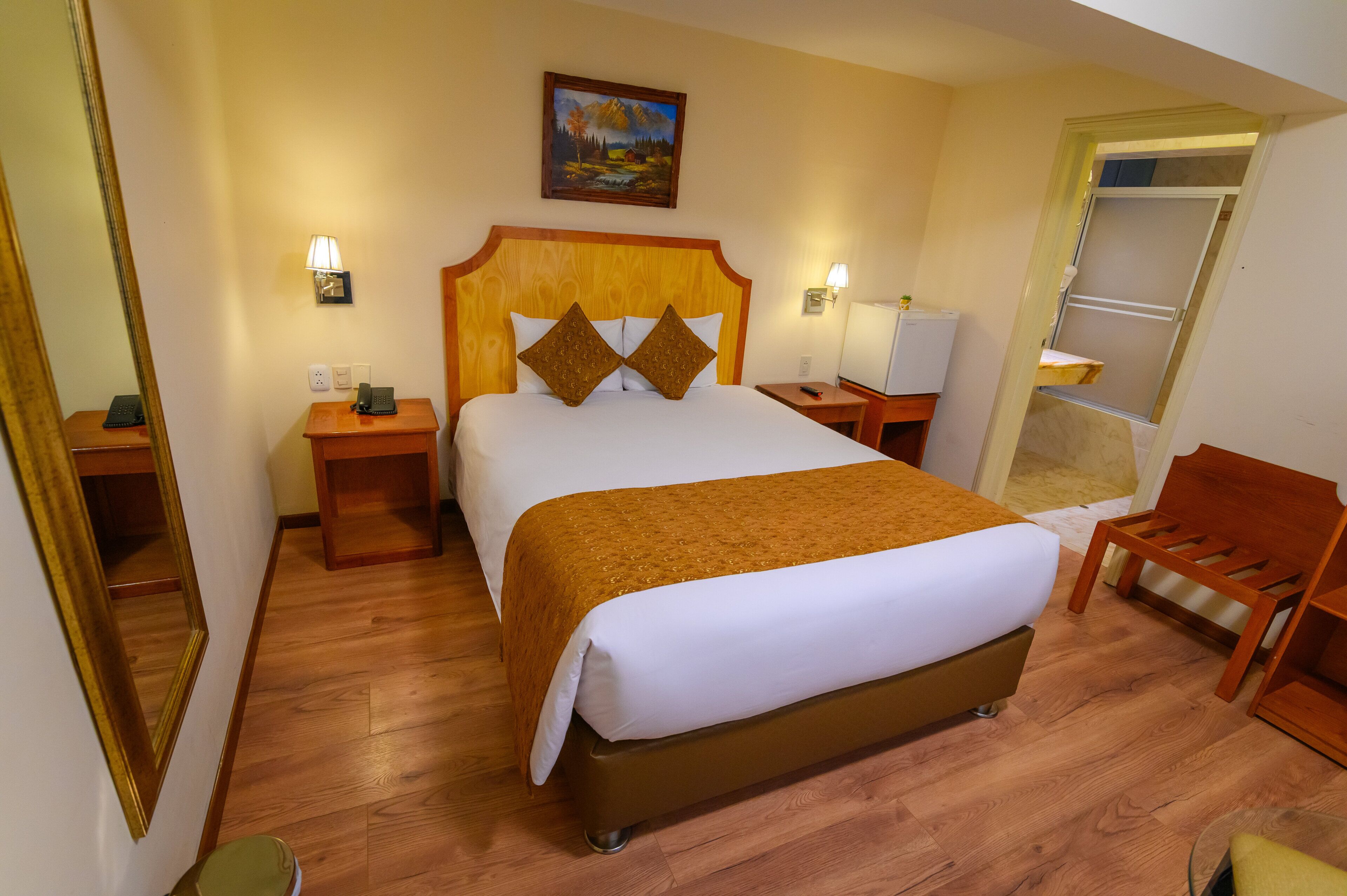 Una habitación de hotel con una cama grande, cabecera de madera, mesitas de noche, una lámpara, un cuadro en la pared y una silla de madera.