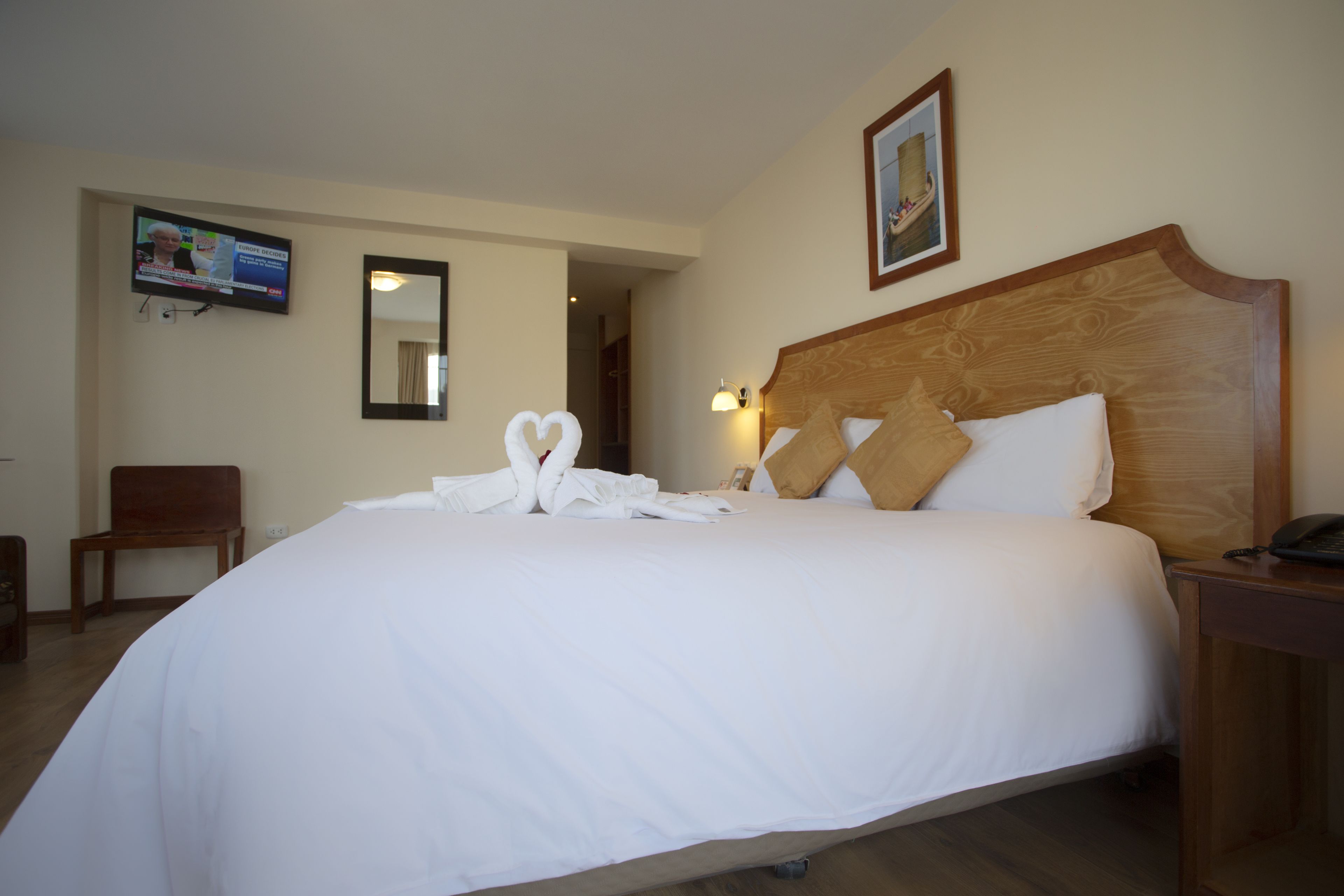 Een hotelkamer met een groot bed, een houten hoofdbord, een nachtkastje met een lamp, een aan de muur bevestigde televisie en een spiegel.