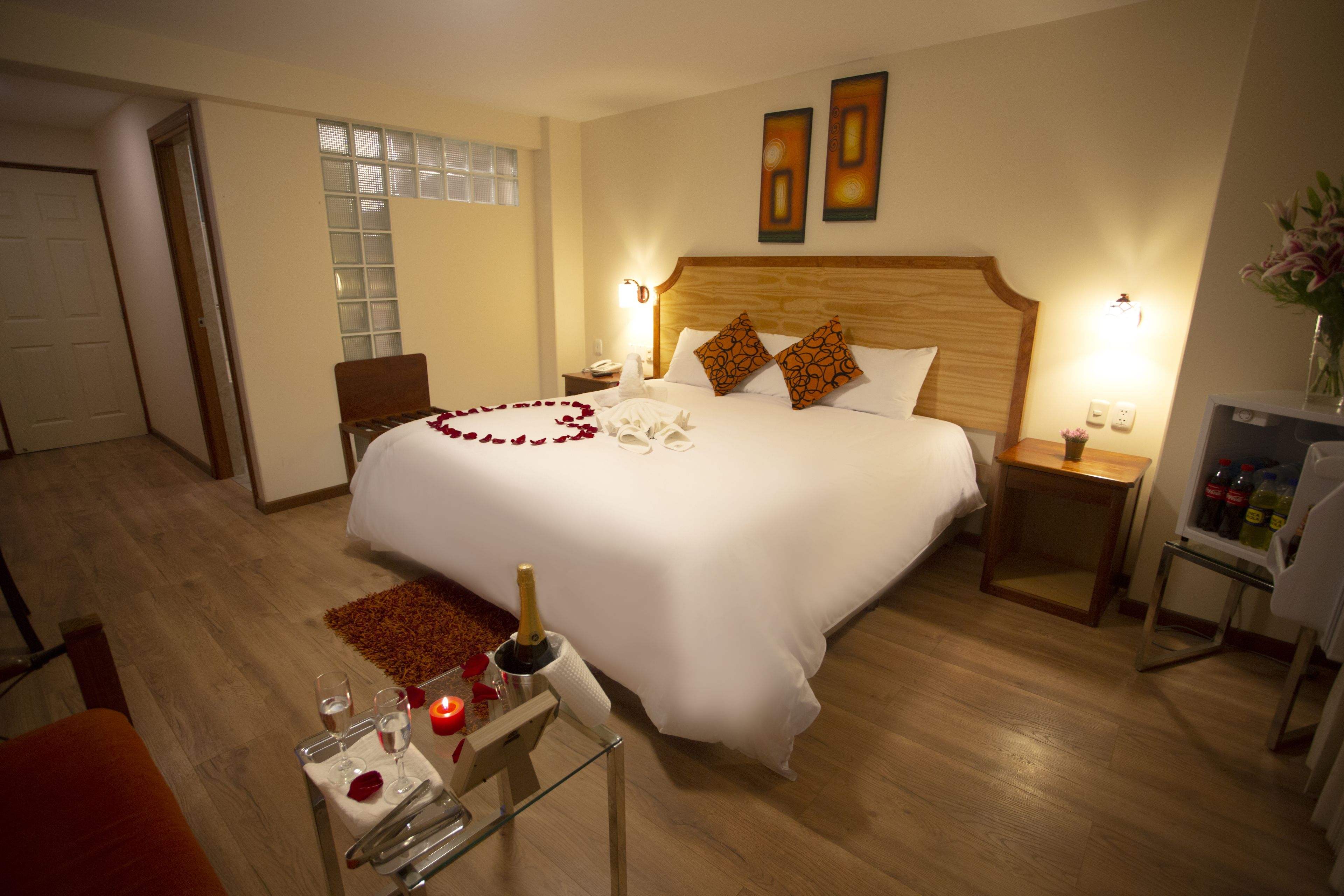 Ein Hotelzimmer mit einem großen Bett, einem hölzernen Kopfteil, zwei Nachttischen mit Lampen, einem kleinen Tisch mit Champagner, Gläsern und einer Rose.