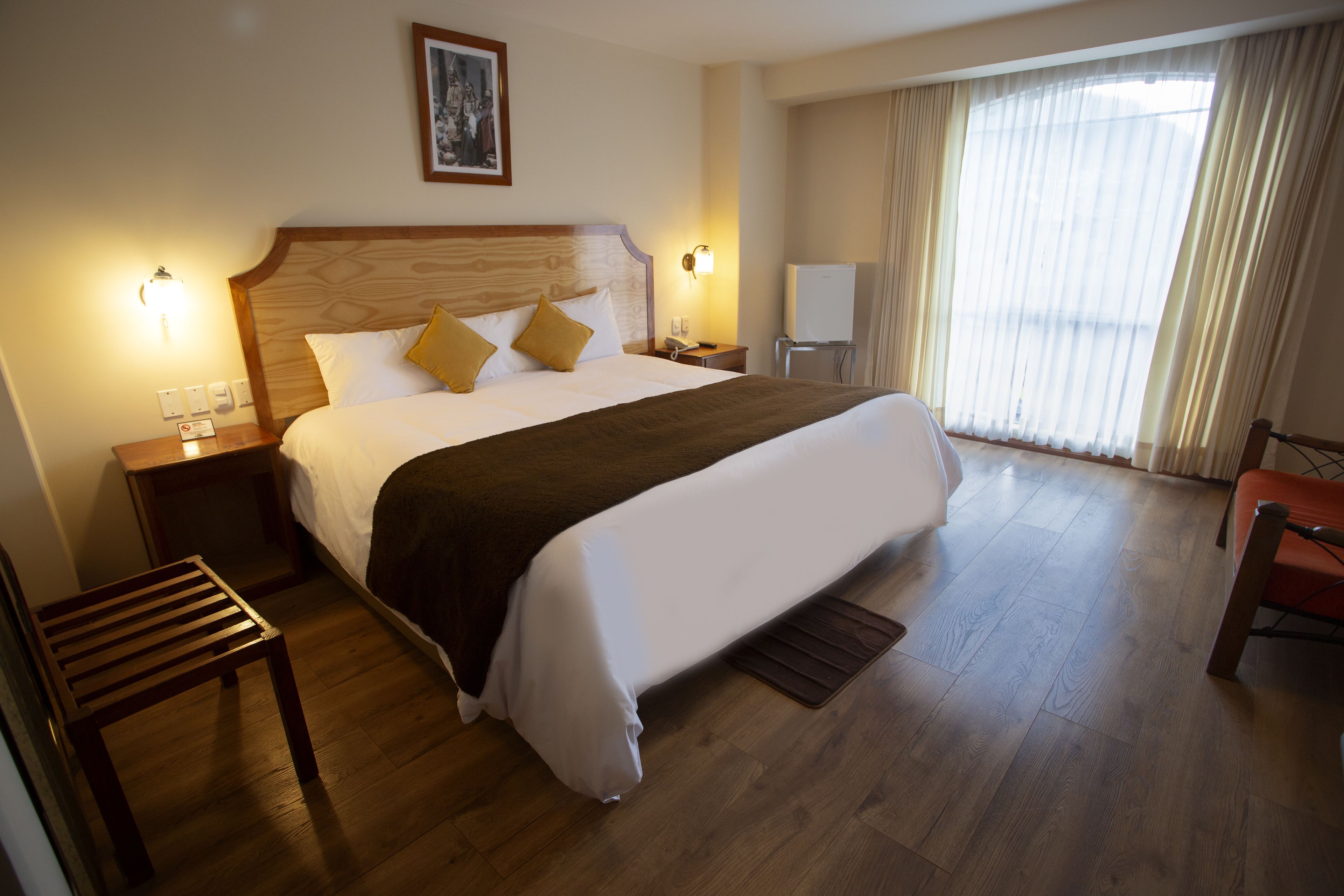 A hotel room with a large bed, wooden floor, a nightstand, a chair, and a window with curtains.