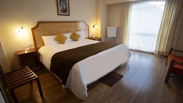 A hotel room with a large bed, wooden floor, a nightstand, a chair, and a window with curtains.