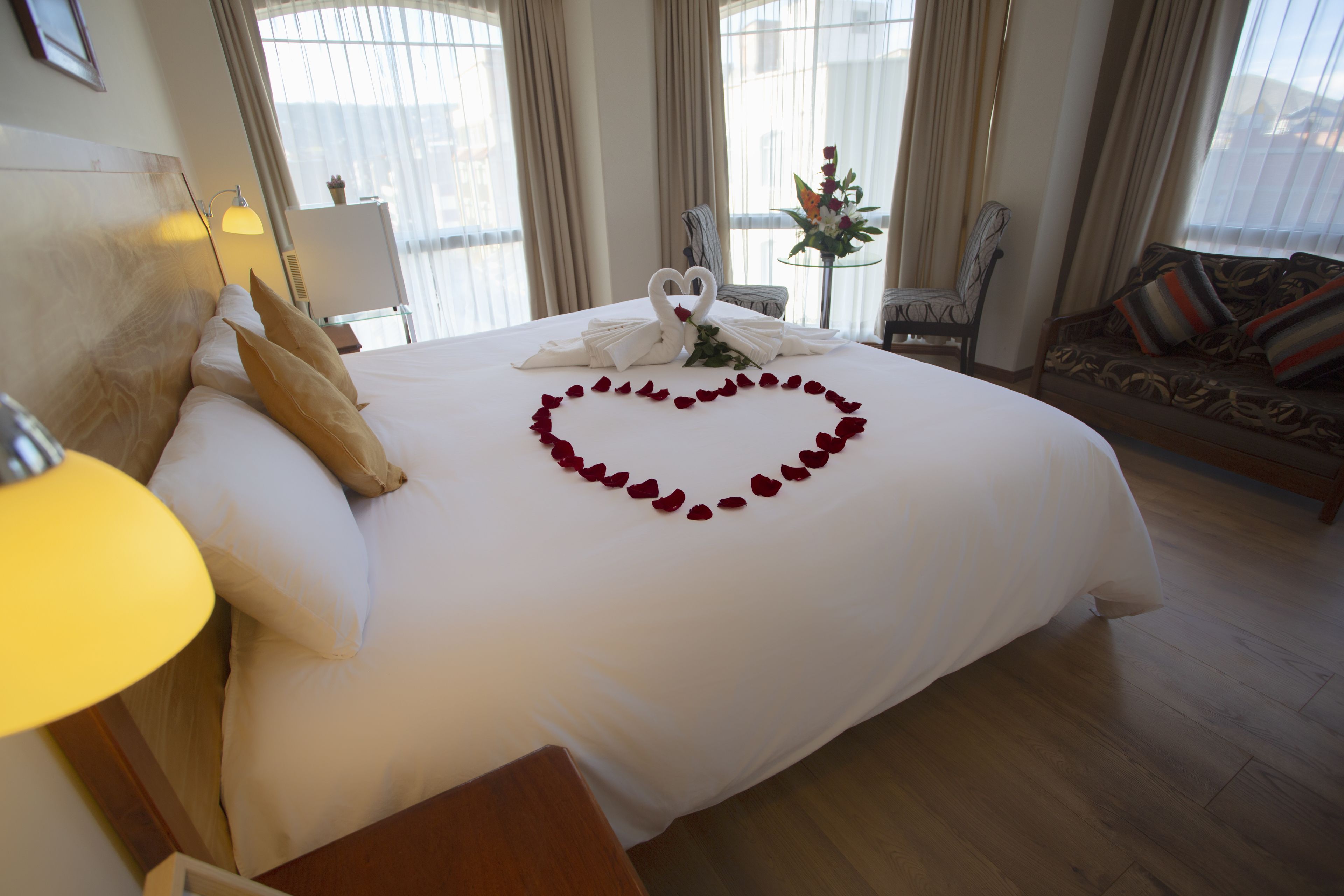 Habitación de hotel con una cama impecable, un arreglo de pétalos de rosa en forma de corazón y una toalla con forma de cisne, y vista a la ciudad a través de amplias ventanas.