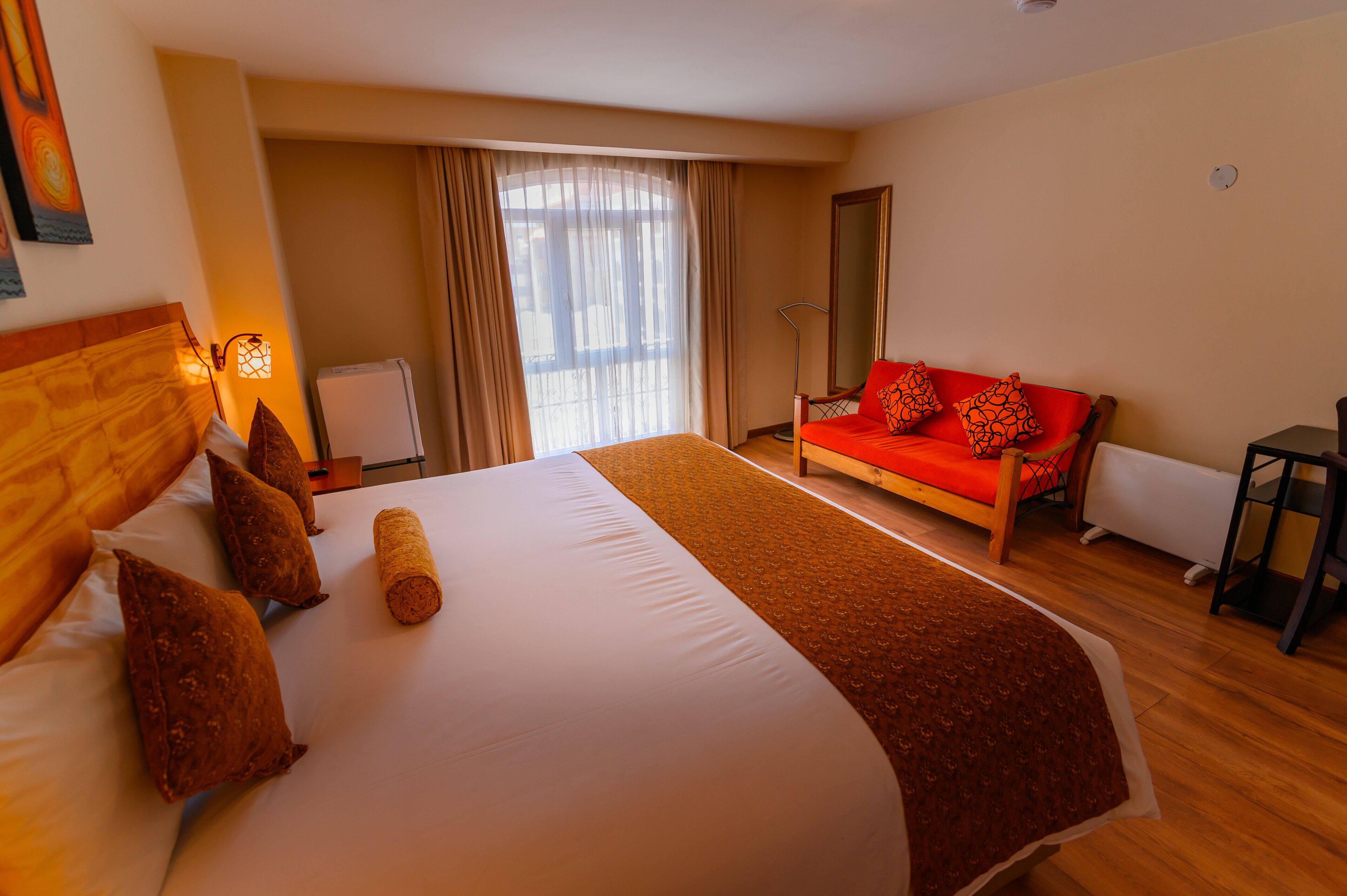 A hotel room with a large bed, a red sofa, a standing lamp, and a view of the outdoors through a window.