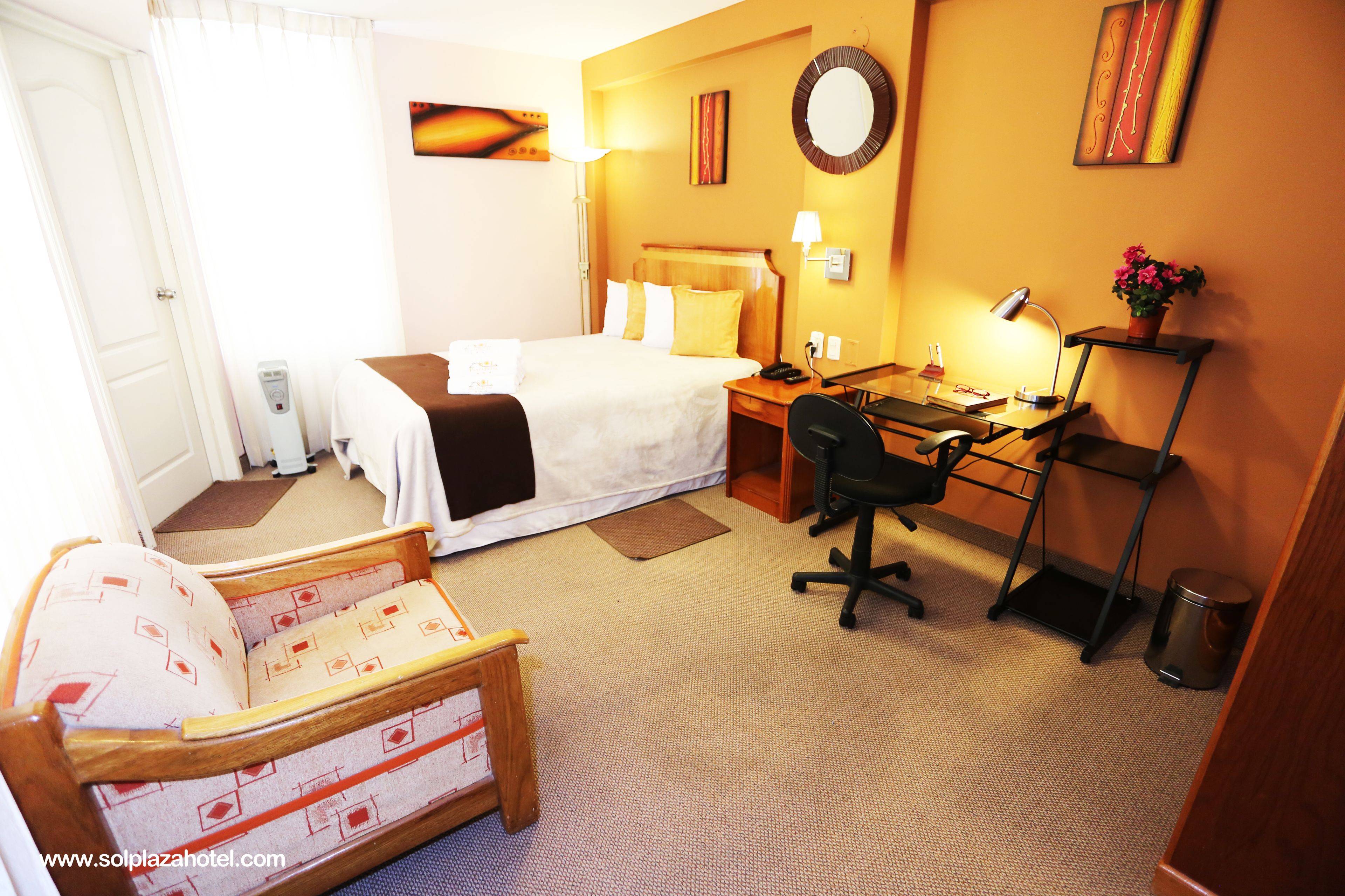 A hotel room with a bed, desk, chair, and a small table with a lamp.