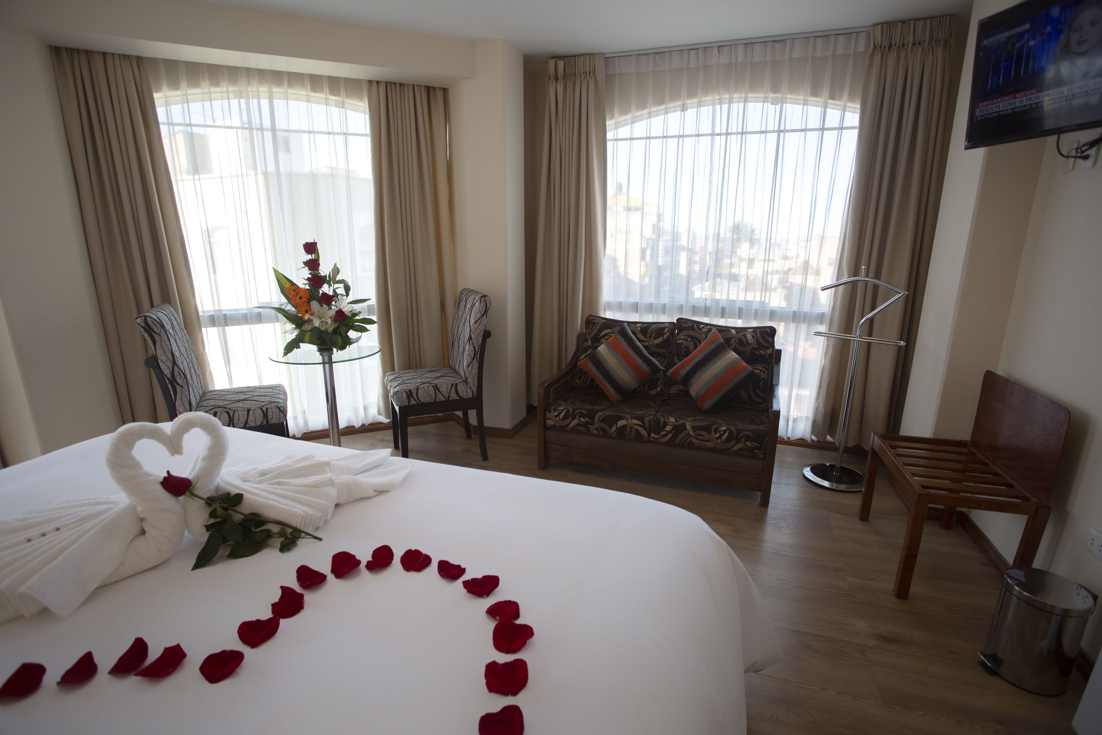 Habitación de hotel con una cama grande, un sofá, una silla, una mesita con un jarrón y un televisor en la pared.
