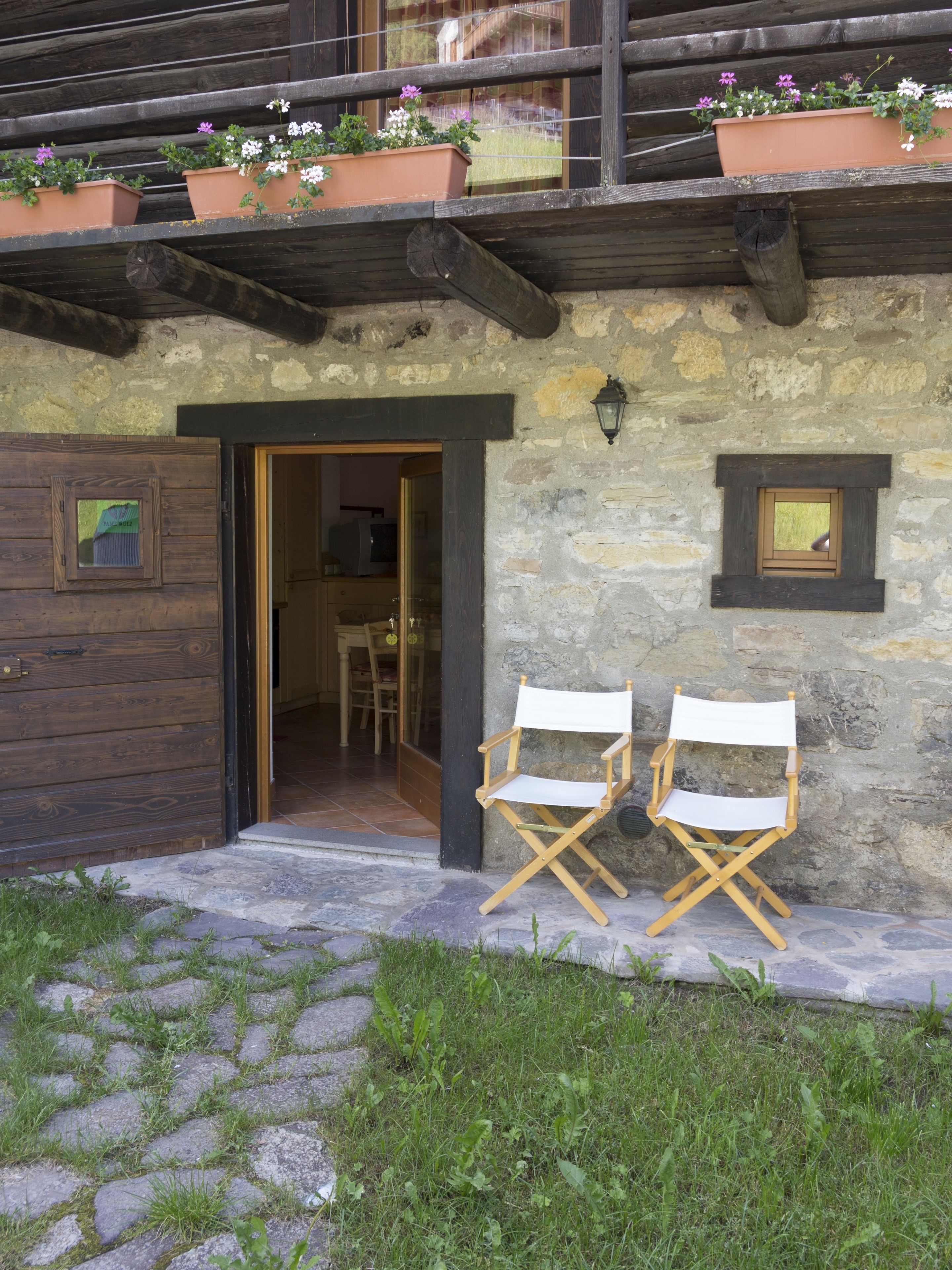 A rustic stone building with a wooden door, two folding chairs, and potted flowers on the balcony.
