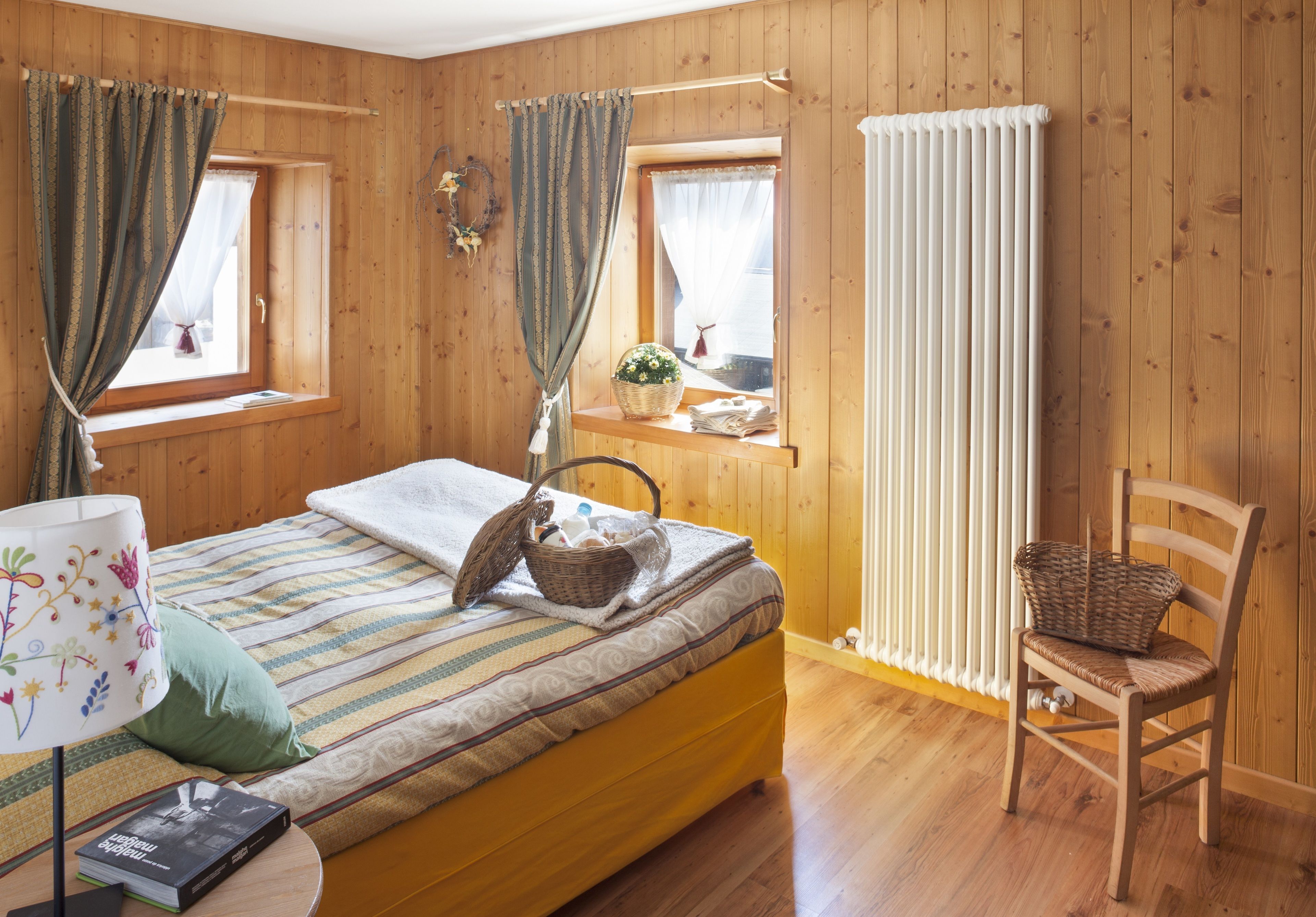 Une pièce confortable avec un lit, une chaise, une fenêtre avec des rideaux, un radiateur et une petite table sur laquelle se trouvent une lampe et un livre.