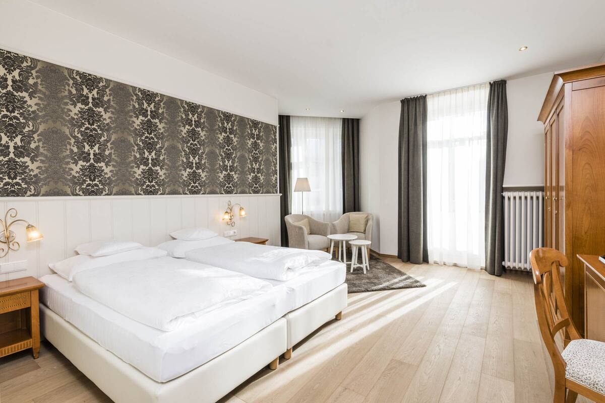 A hotel room with two beds, a desk, a chair, a lamp, and a large window with curtains.