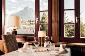 A dining area with a table set for two, a window with a mountain view, and a lamp on the table. - Hotel Villa Kastelruth (Castelrotto)