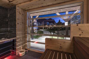 A modern interior with a wooden bench, a stone fireplace, and a large window offering a view of a mountain and a pathway.