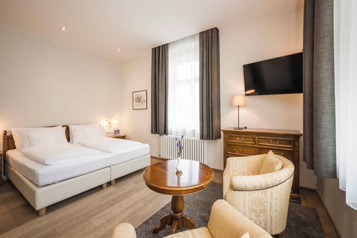 A hotel room with a bed, a round wooden table, a chair, a dresser, a TV, and a window with curtains.