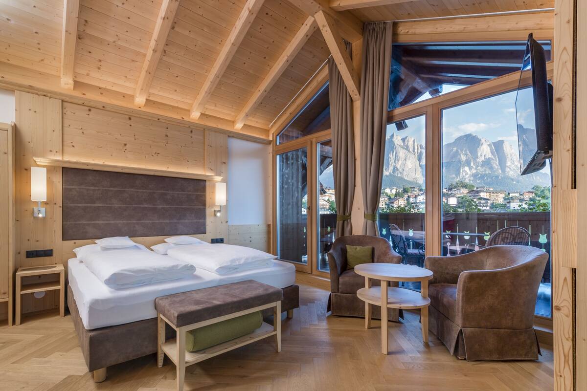 A wooden room with a bed, a table, and a chair. There is a window with a view of mountains.