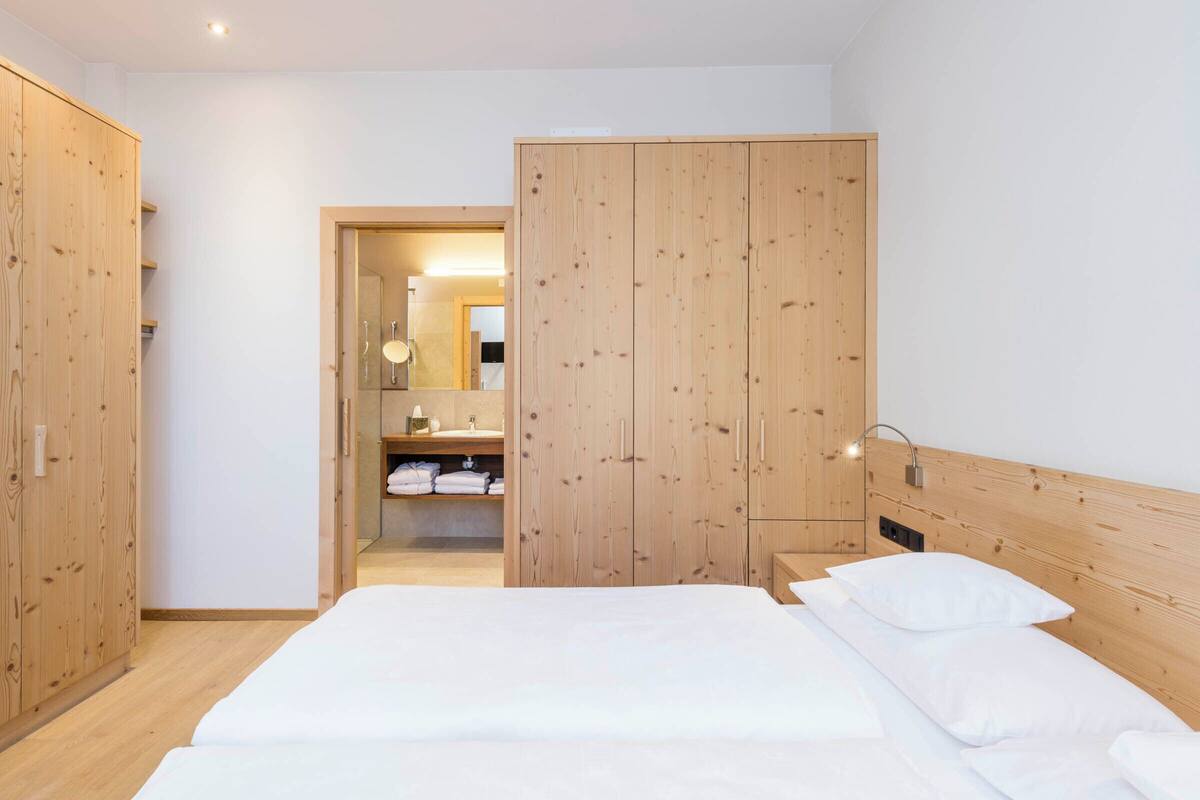 A bedroom with a wooden wardrobe, a bed with white bedding, and a nightstand with a lamp.