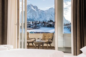 A balcony with a wicker chair, a small table, and a view of snow-covered mountains and a village. - Hotel Villa Kastelruth (Castelrotto)