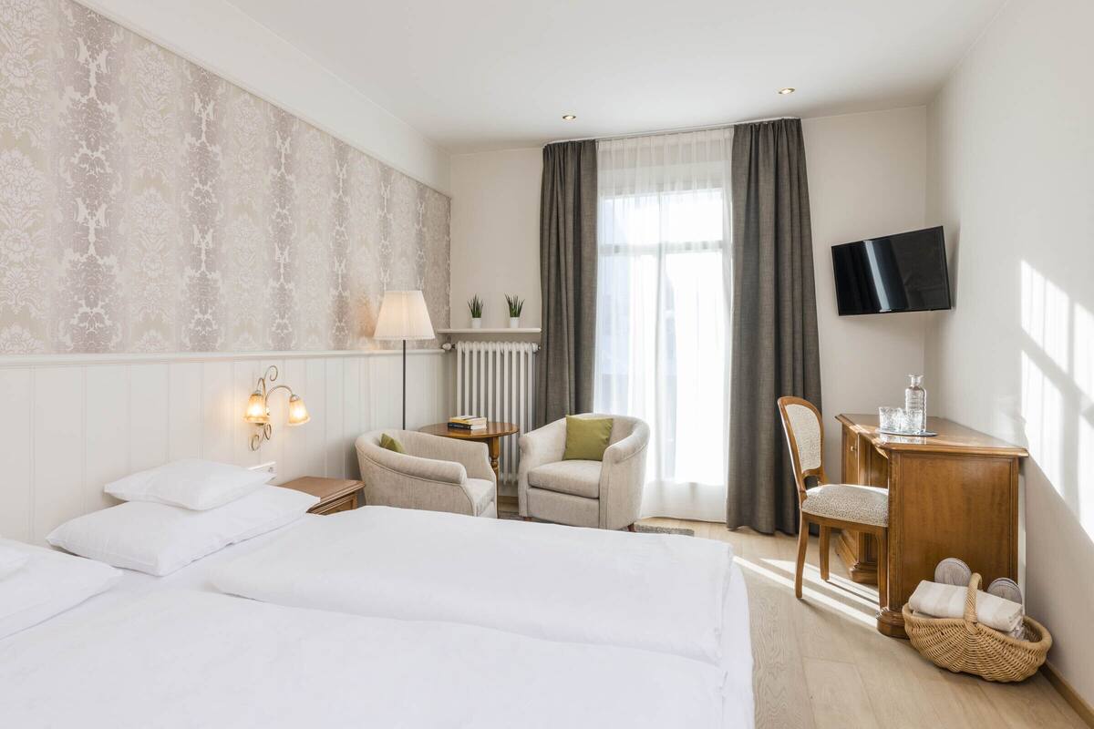 A hotel room with two beds, a desk with a chair, a TV, and a window with curtains.