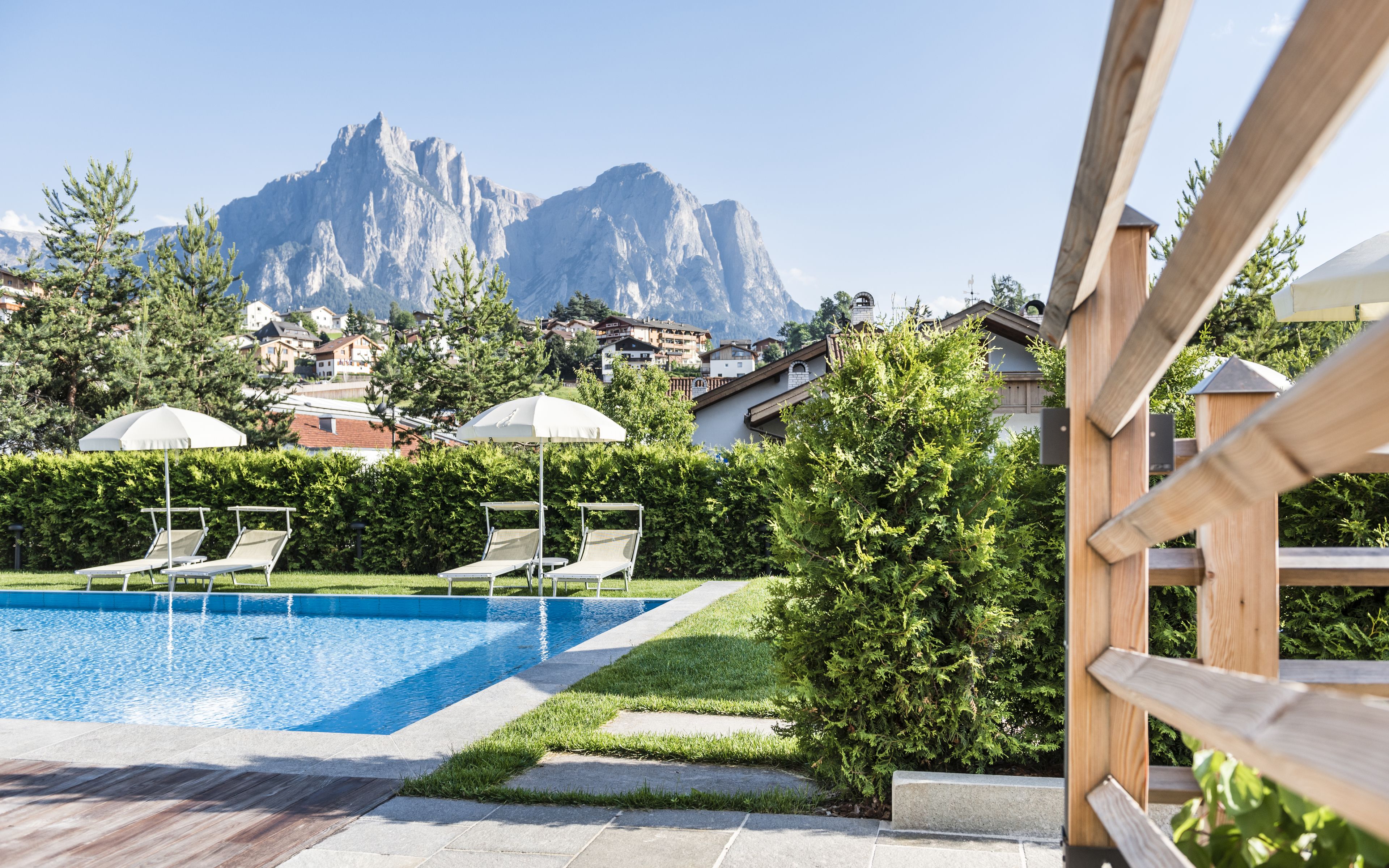 A swimming pool with lounge chairs and umbrellas, surrounded by greenery and buildings, with a mountain in the background.