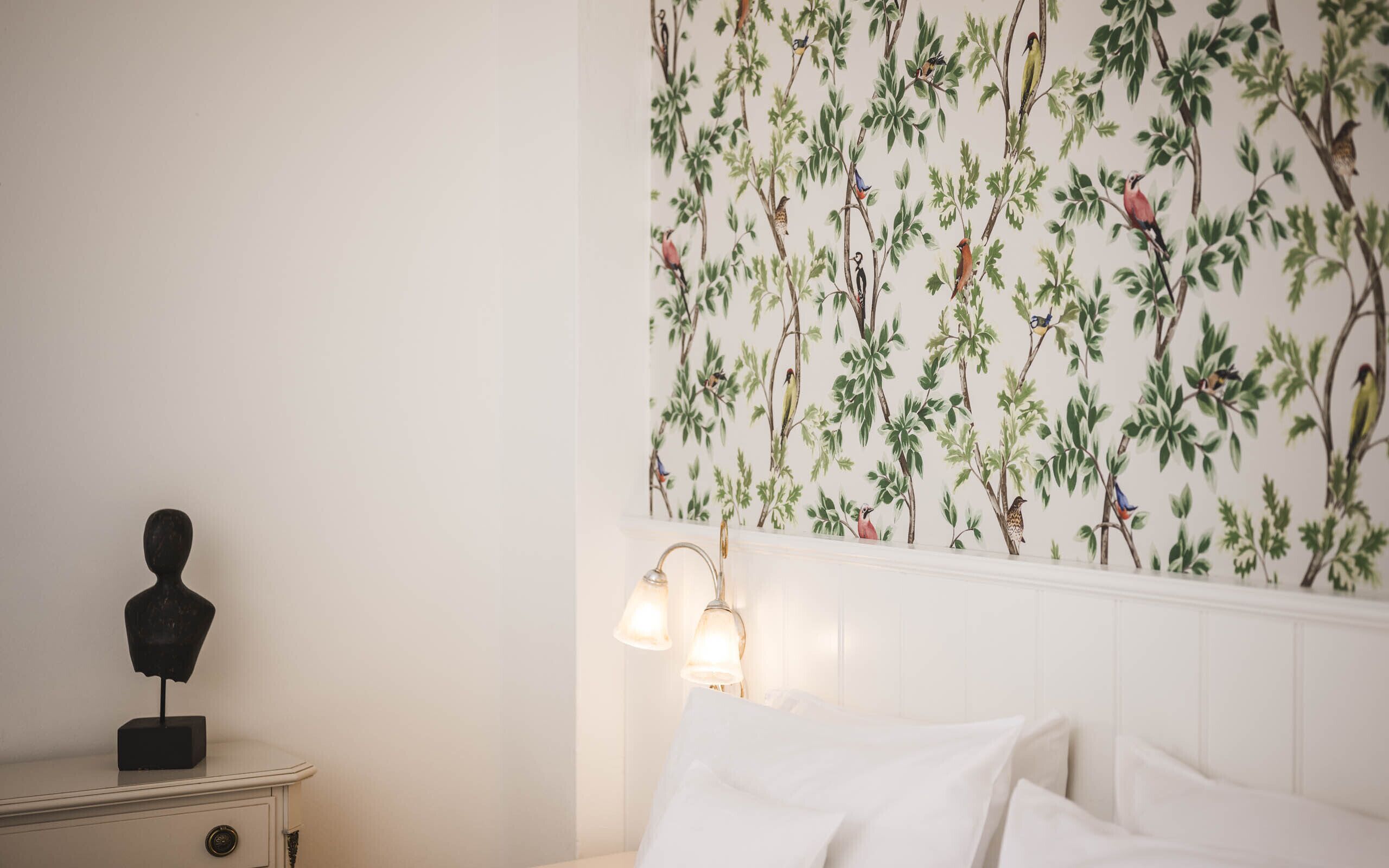 A bedroom with a white headboard, a nightstand with a bust sculpture, and a wall with a bird-pattern wallpaper.