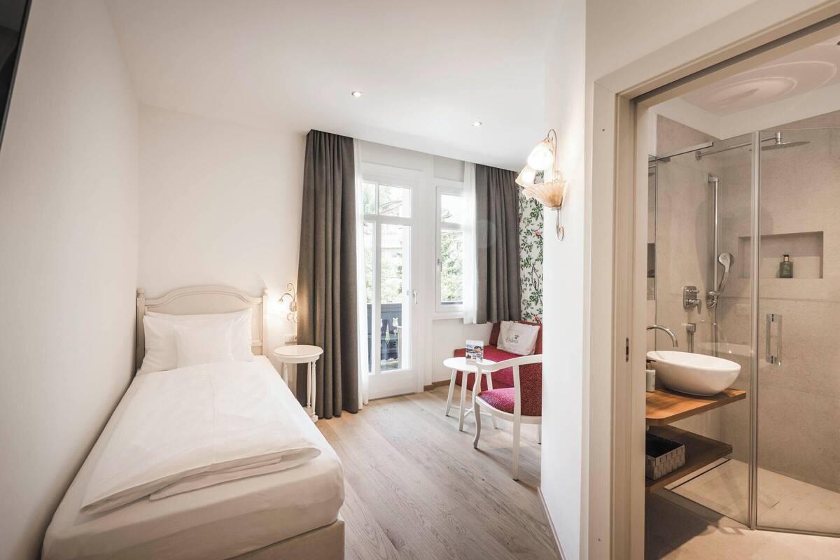 A bedroom with a bed, bedside table, and a small table with a chair. There is a bathroom with a shower and a sink.