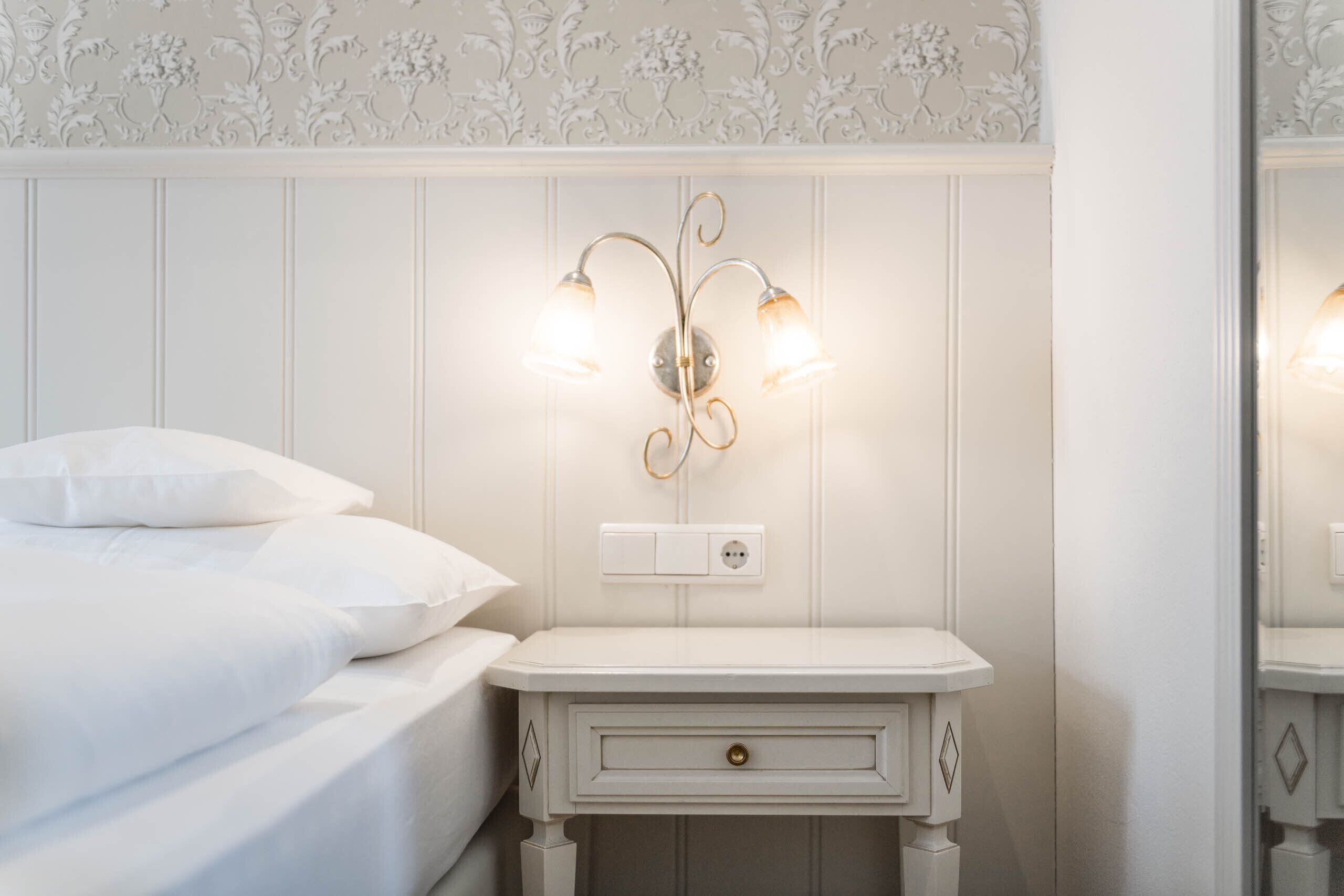 A neatly made bed with white linens and pillows, a white bedside table with a drawer, and a wall-mounted light fixture with three bulbs.