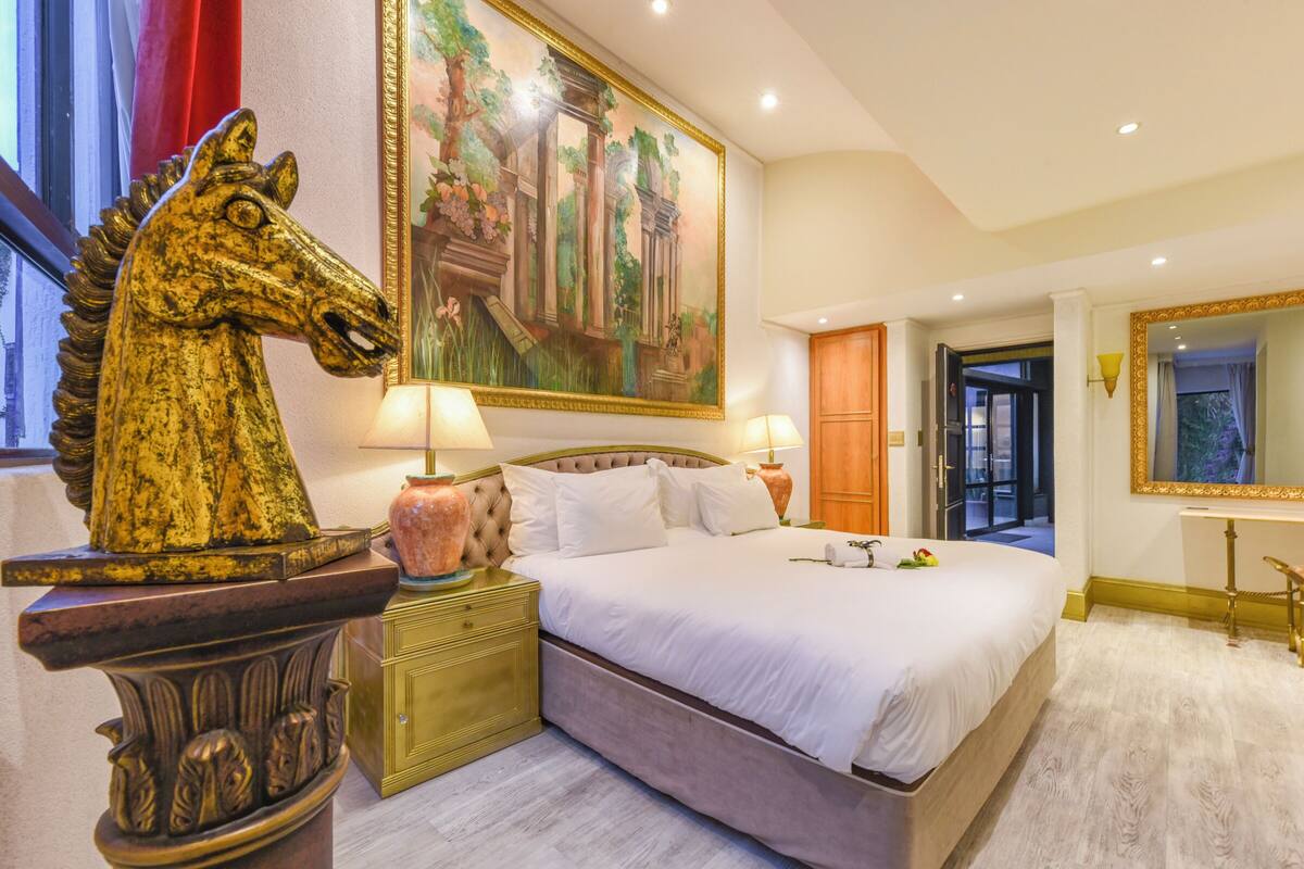 Un dormitorio con una cama grande, dos mesitas de noche con lámparas, una estatua decorativa de un caballo, un cuadro grande enmarcado y un espejo.