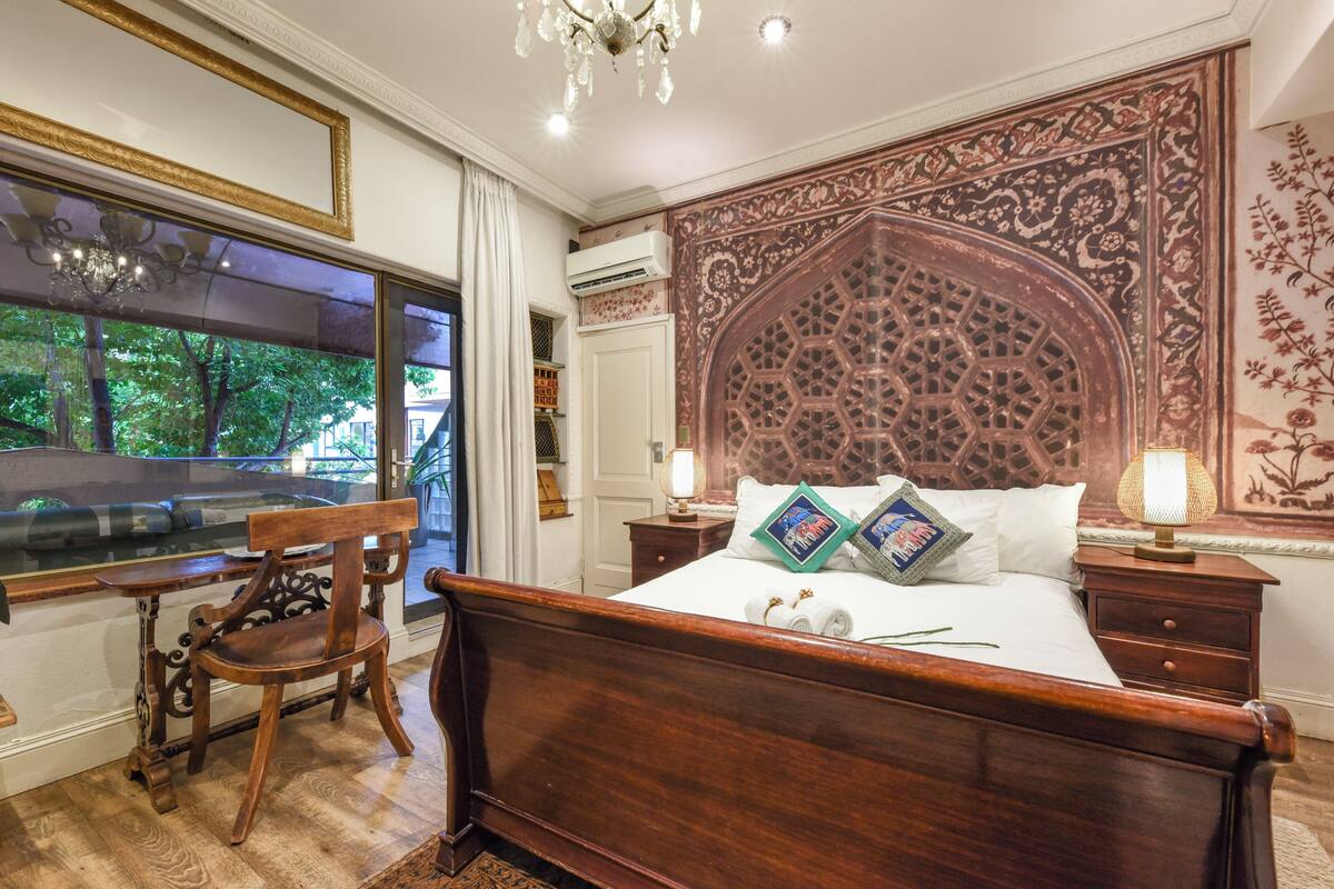 Un dormitorio con una cama de madera, un escritorio, una silla, un ventanal grande y un panel decorativo en la pared.